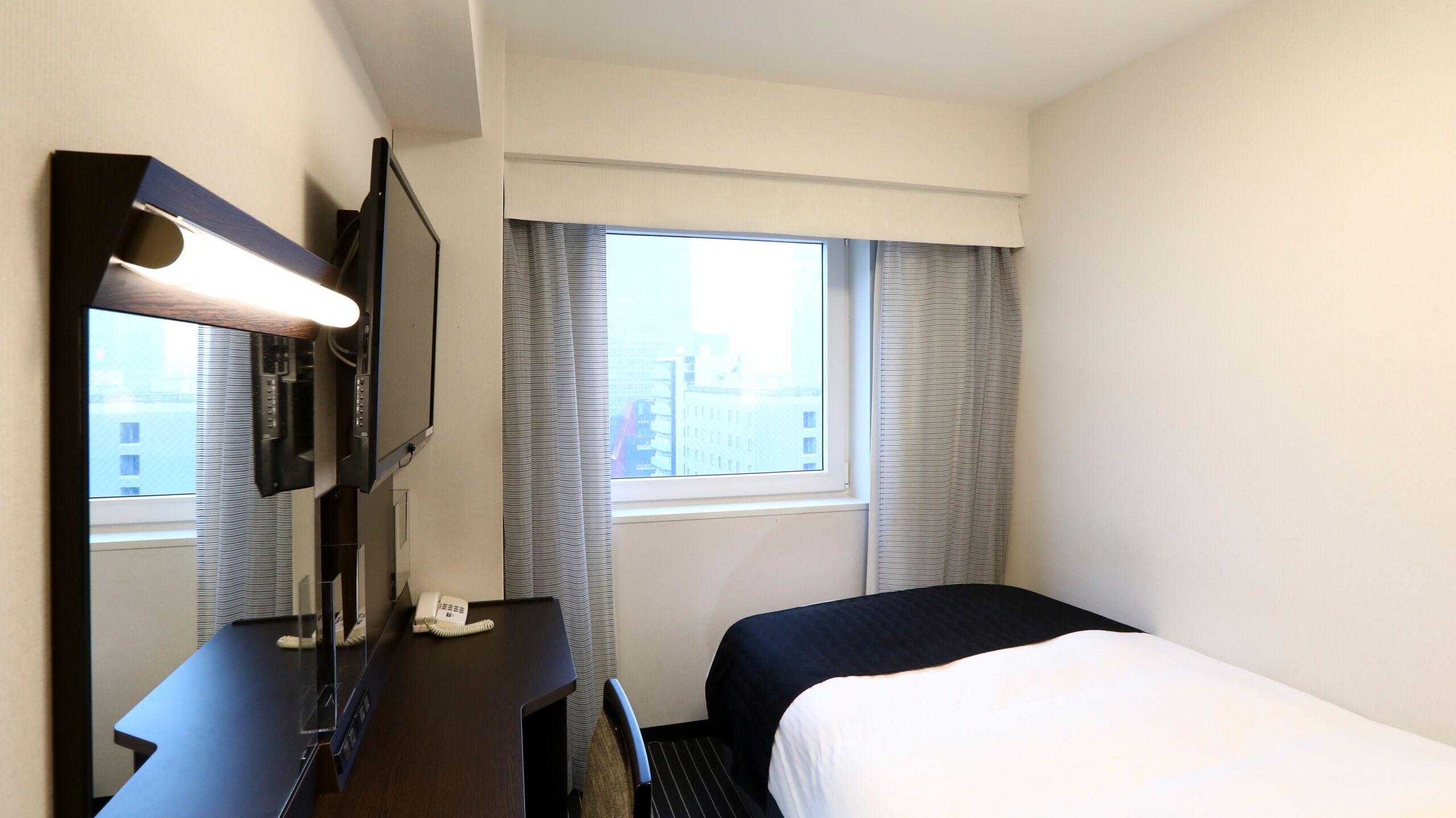 A hotel room with a bed, a desk with a telephone, a television, and a window with curtains.
