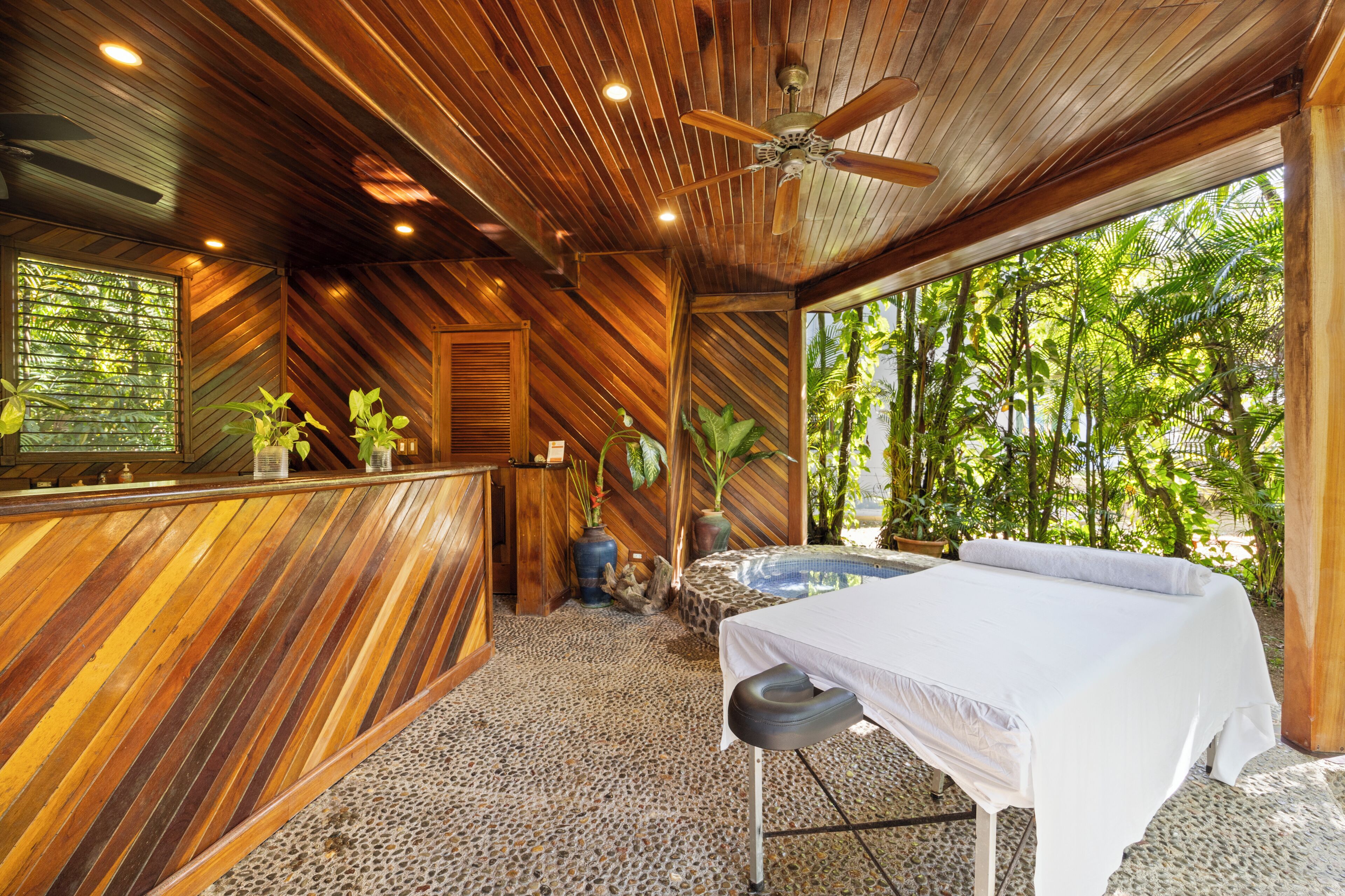 Een spa-ruimte met houten lambrisering, een receptiebalie, een massagetafel en uitzicht op weelderige begroeiing.