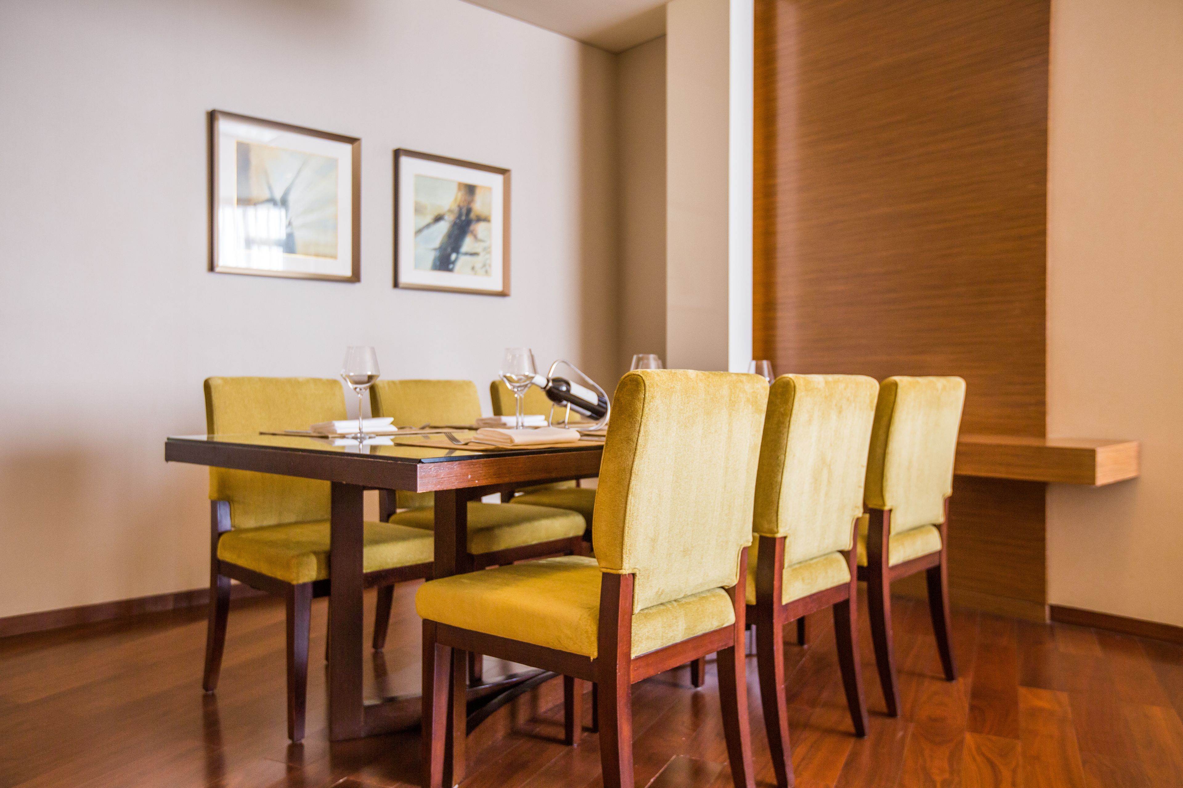 A dining area with a wooden table and yellow chairs, framed pictures on the wall, and a wooden panel on the right.