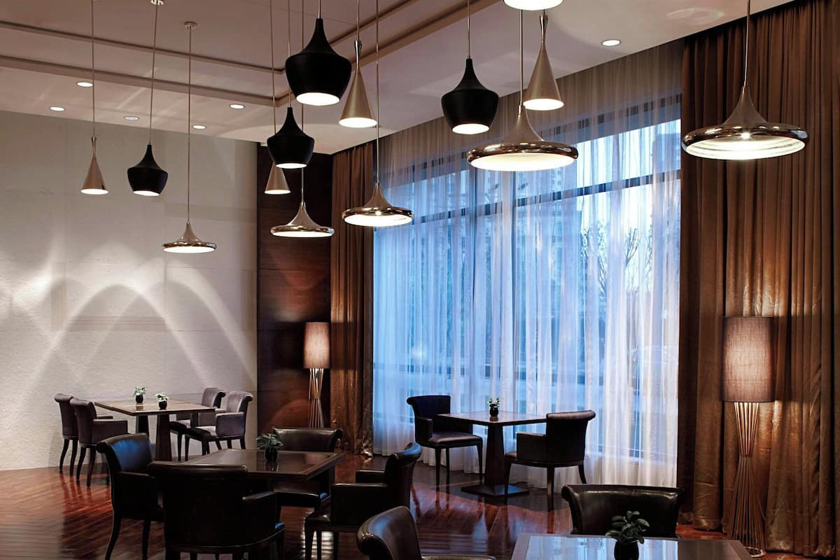 Un comedor amplio con varias mesas y sillas, ventanas grandes con cortinas y lámparas colgantes.
