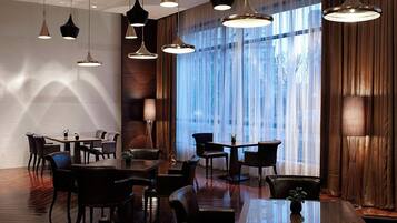 A spacious dining area with multiple tables and chairs, large windows with curtains, and hanging pendant lights.