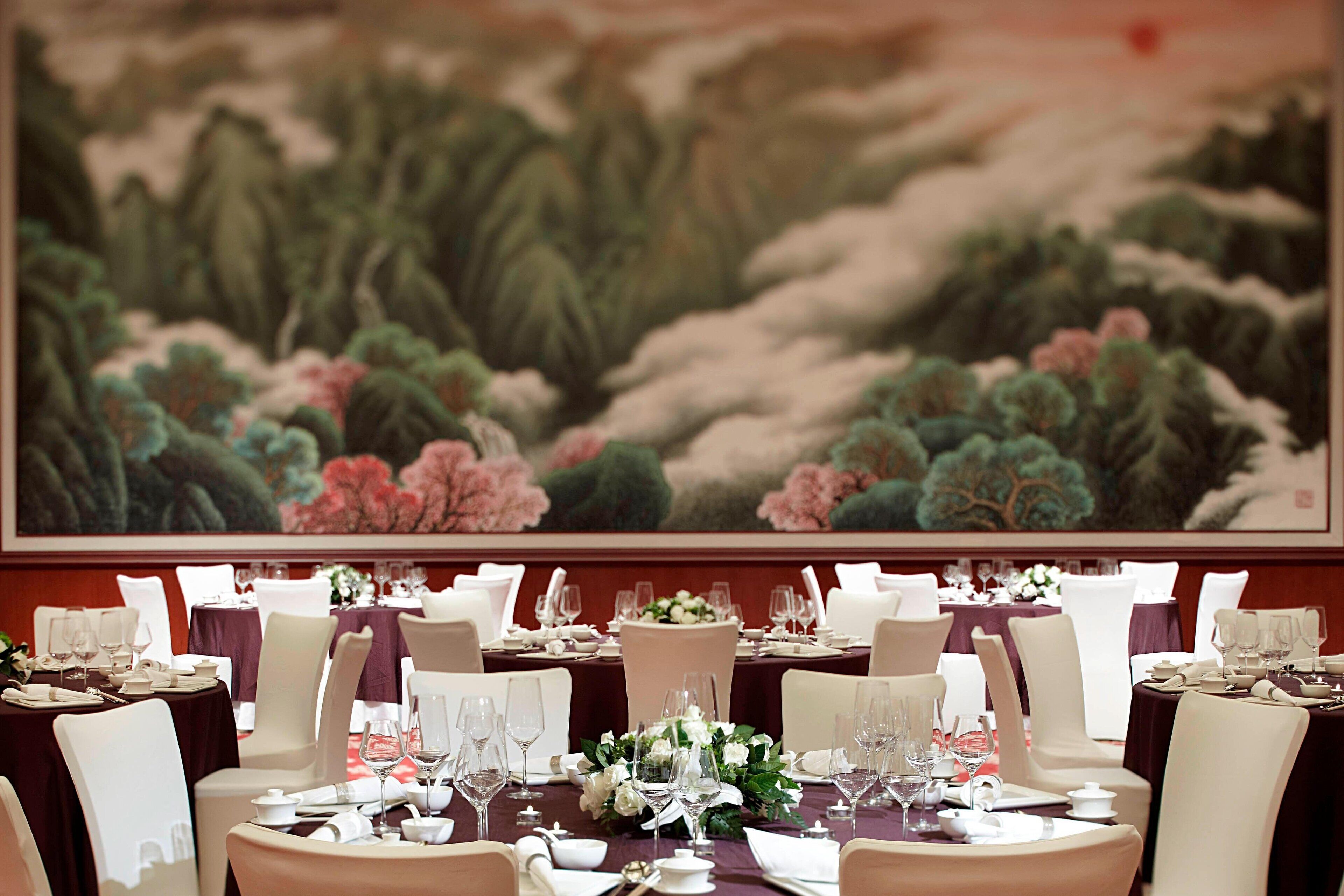Un comedor formal con mesas dispuestas para una comida, un cuadro grande en la pared y vista a un jardín.
