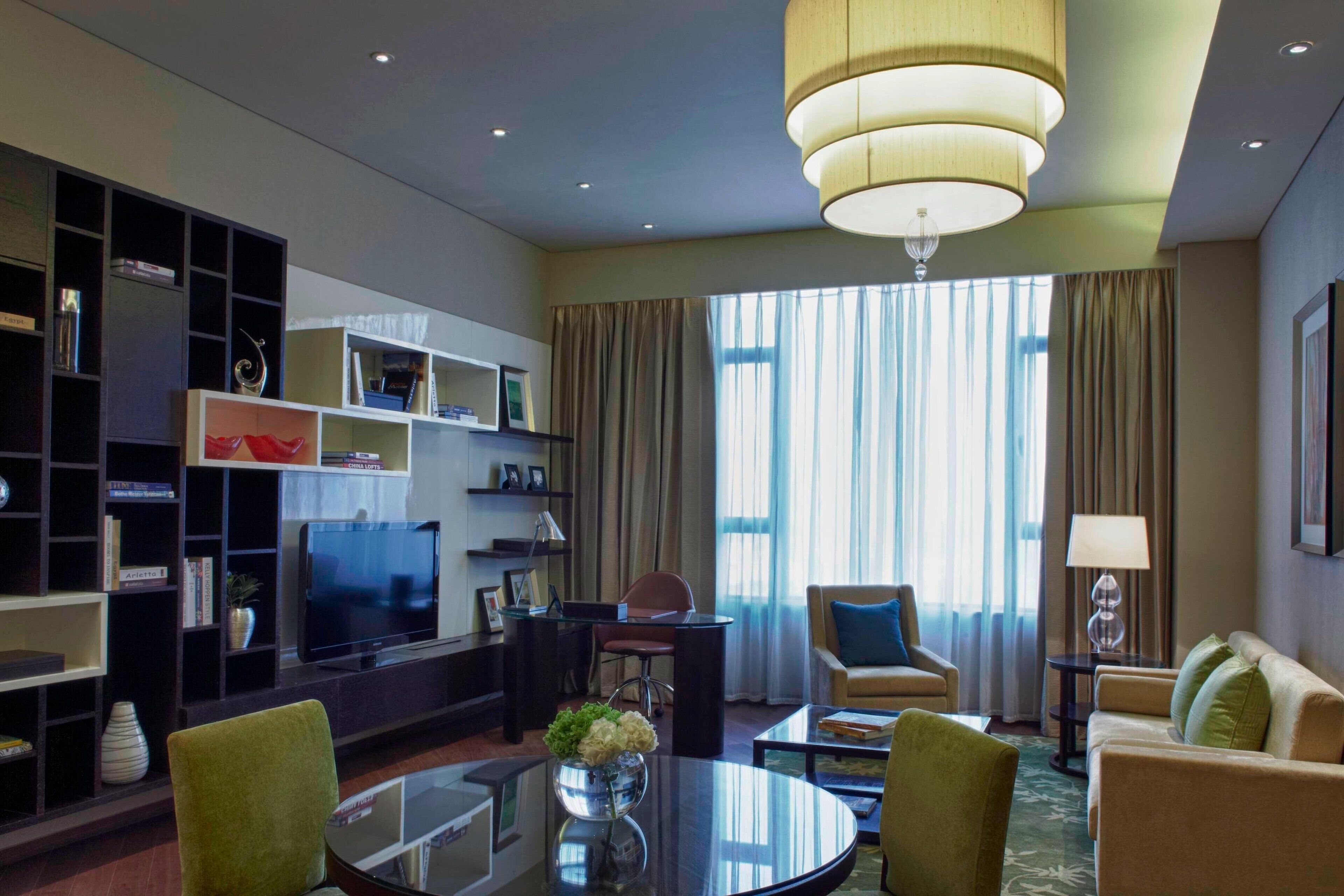 Una habitación de hotel moderna con una televisión de pantalla plana, una mesa de centro redonda, un sofá, un escritorio con una silla, una estantería y un ventanal con cortinas.
