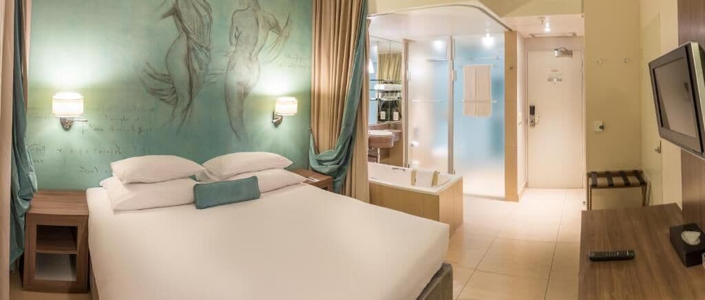 Habitación de hotel con cama, lámparas de noche, televisión, baño con ducha y espejo.