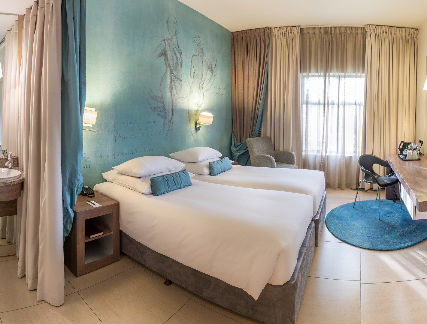 A hotel room with two beds, a desk, a chair, a bathroom, and a large window.
