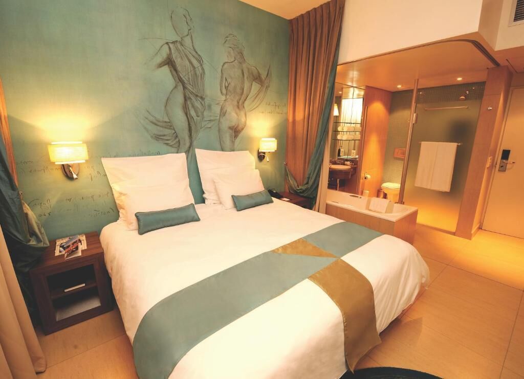 A hotel room with a large bed, two bedside lamps, a nightstand, a bathroom visible through a glass door, and a mural on the wall.