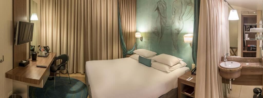 Habitación de hotel con cama, escritorio, silla, televisor, lámpara de escritorio, luz en la pared, baño con lavamanos y cabina de ducha.