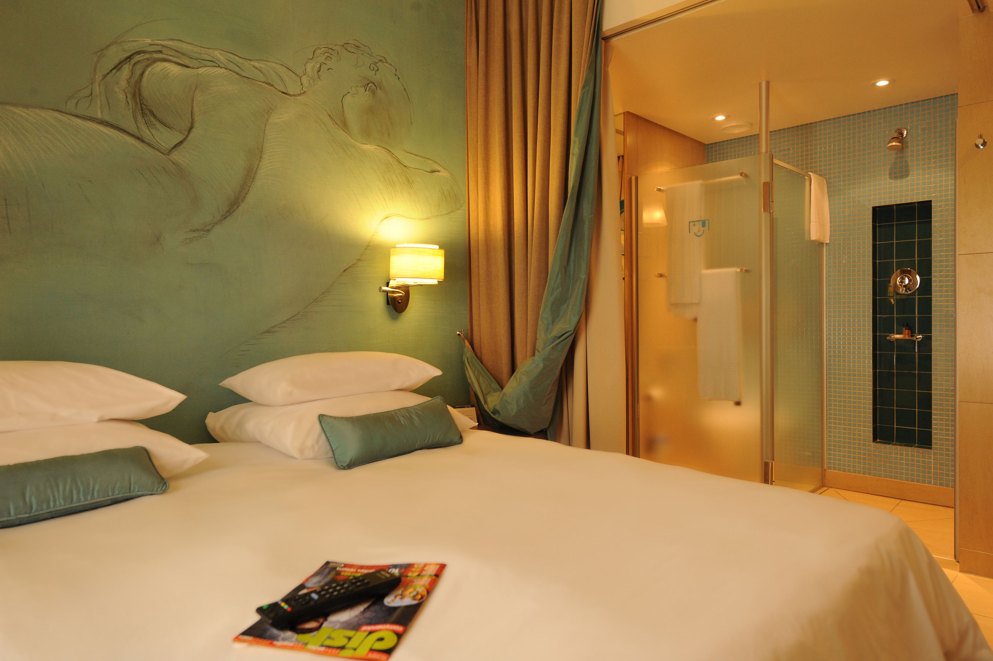 A hotel room with a bed, pillows, a wall-mounted lamp, a shower area, and a tiled wall.