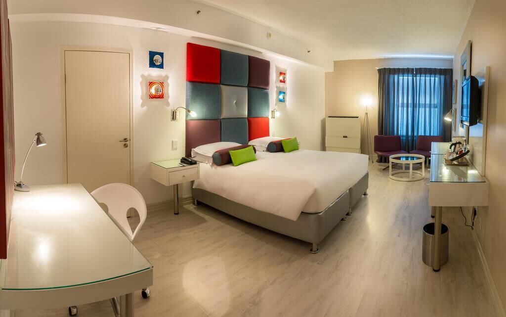 A hotel room with a bed, bedside tables, a desk, a chair, a TV, and a window with curtains.