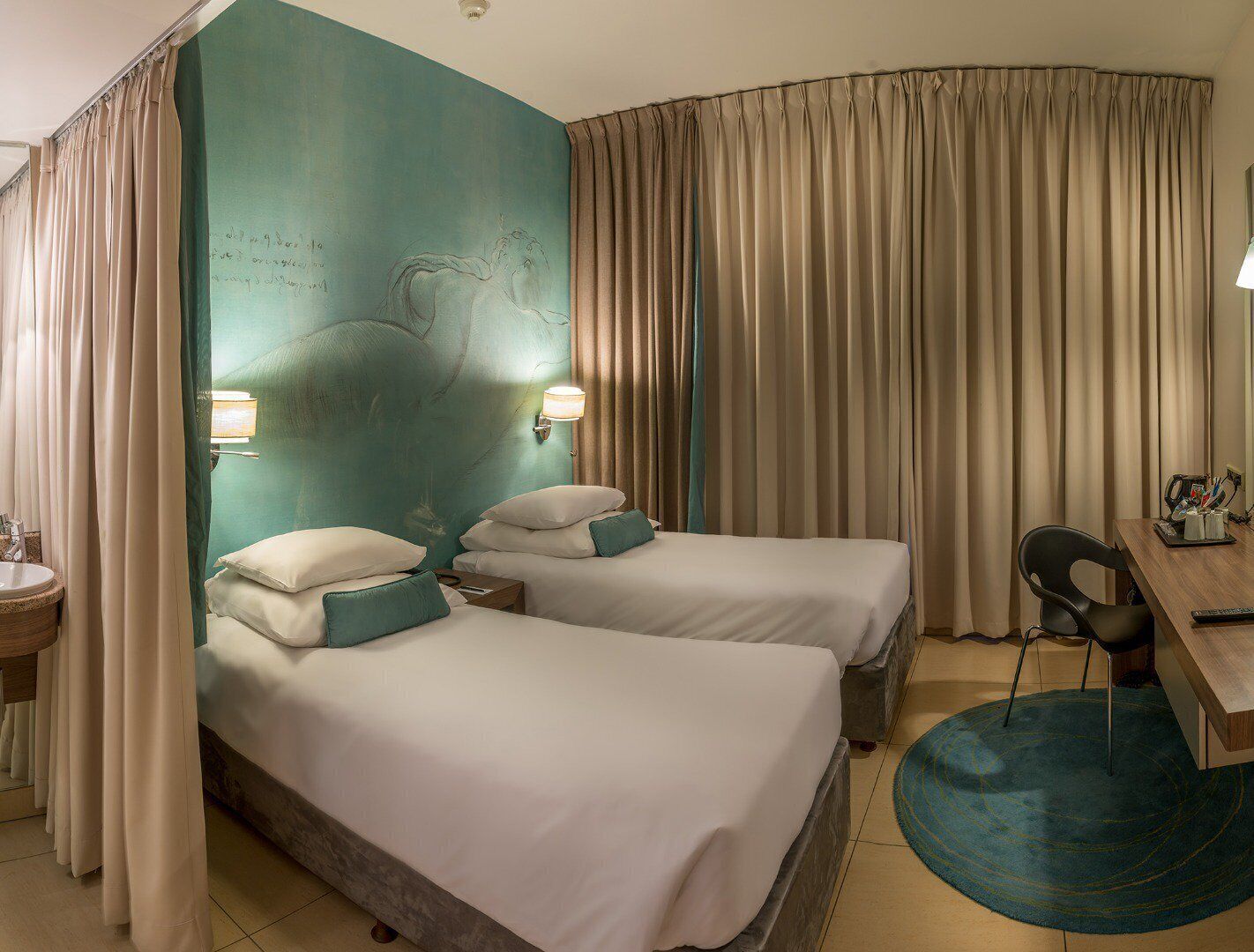 A hotel room with two beds, a desk, a chair, a lamp, and a bathroom with a sink and mirror.