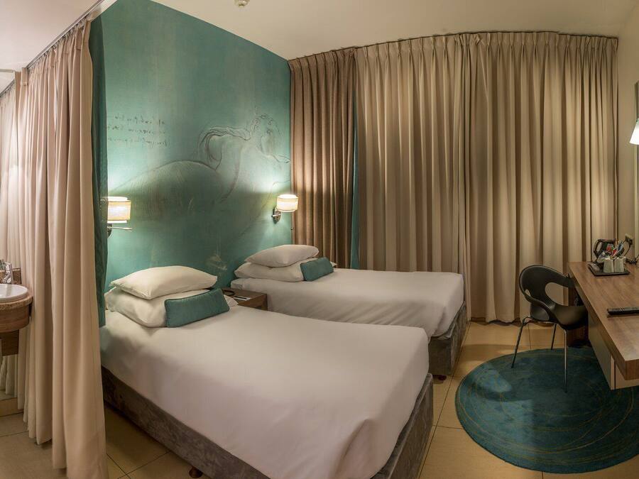 A hotel room with two beds, a desk, a chair, a lamp, and a bathroom with a sink and mirror.