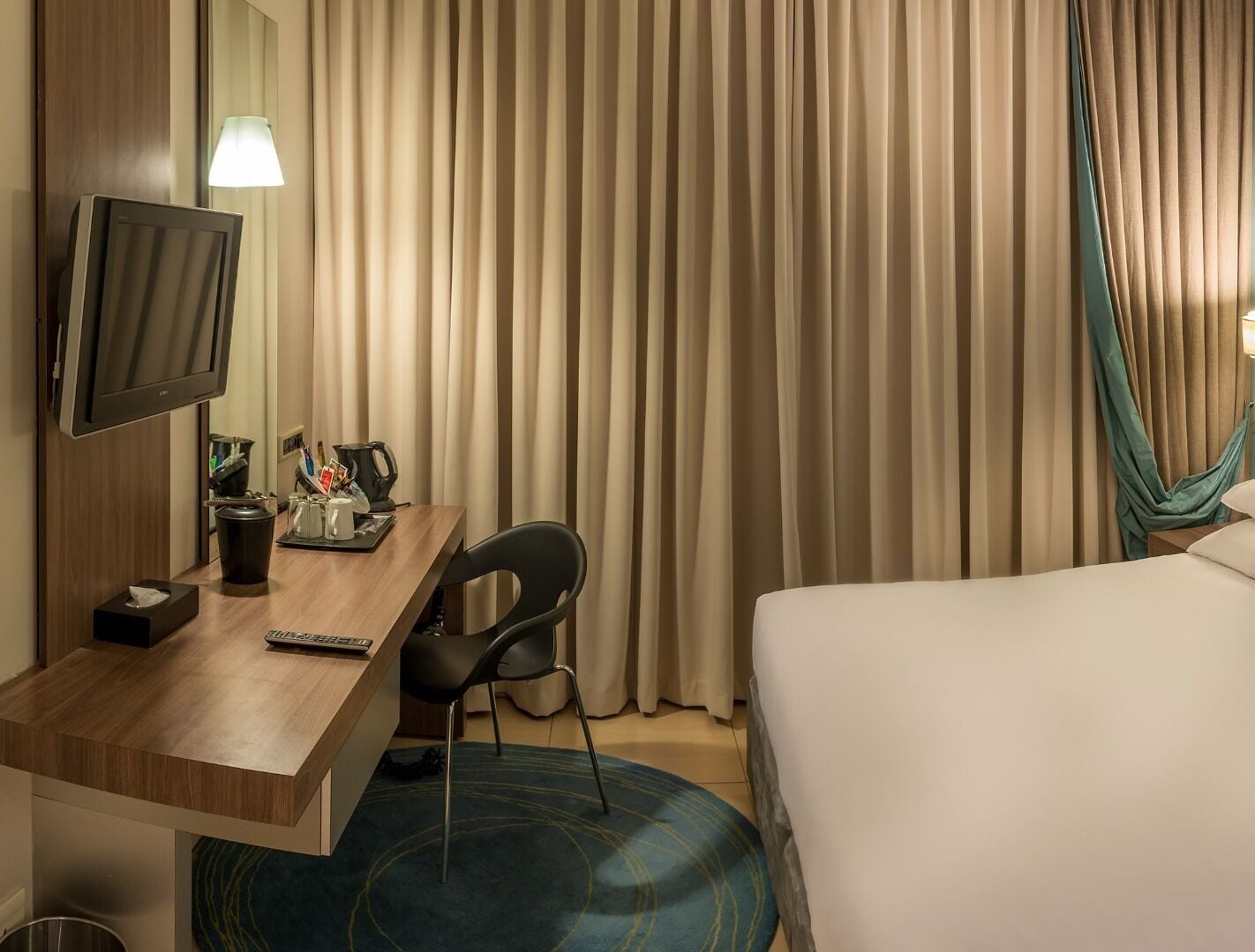 A hotel room with a bed, a desk with a TV, a chair, a coffee maker, and a trash bin.