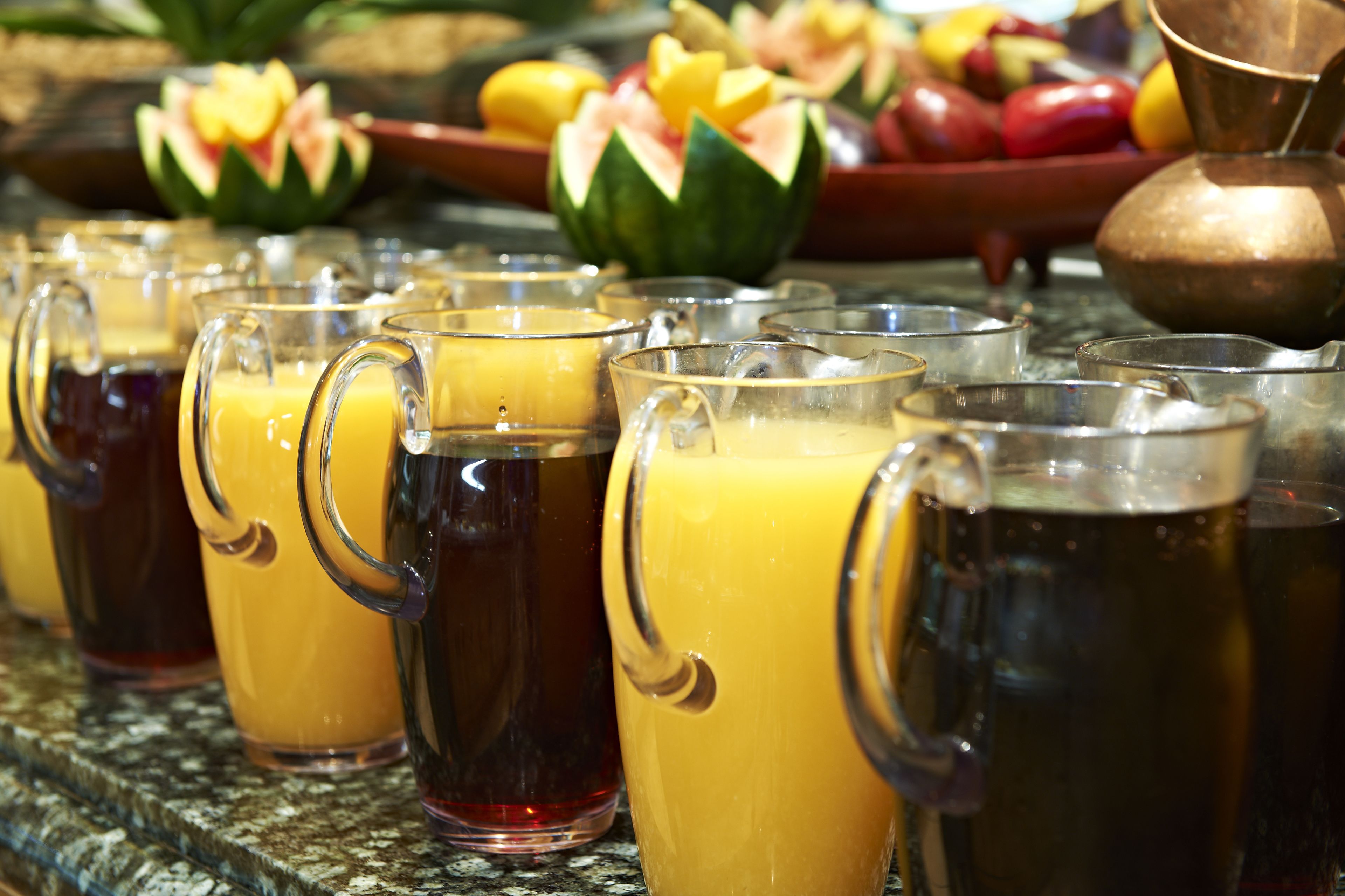 Una hilera de tazas de vidrio transparentes con diversas bebidas, una olla de cobre y un bol con frutas cortadas al fondo.