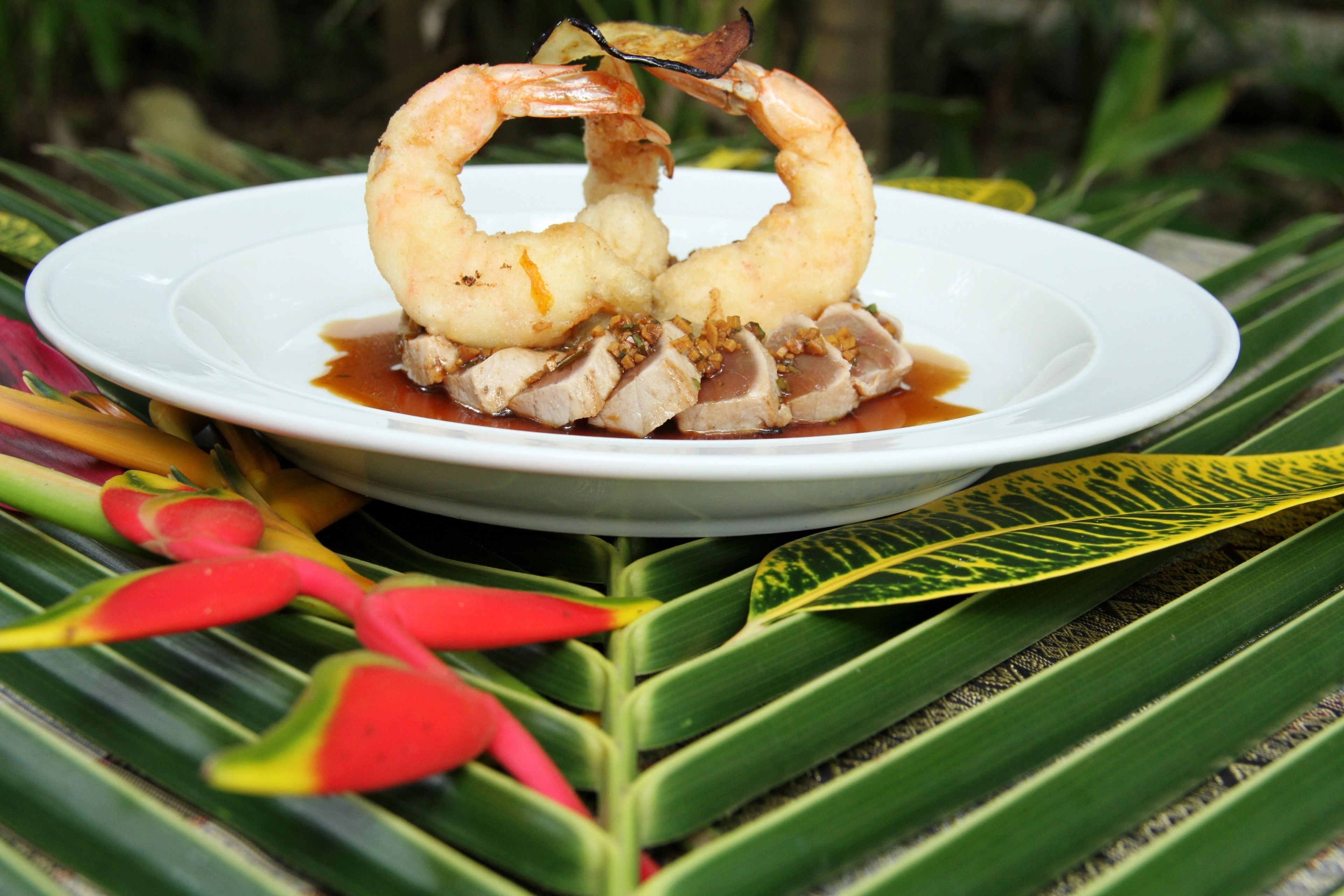 A plate of food with shrimp and meat on a white plate, surrounded by green leaves and red flowers.
