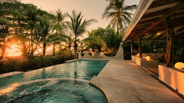 A swimming pool with a curved edge, surrounded by palm trees and a covered seating area with cushions. - Hotel Tropico Latino (Cóbano)