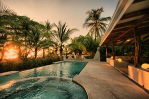 A swimming pool with a curved edge, surrounded by palm trees and a covered seating area with cushions. - Hotel Tropico Latino (Cóbano)