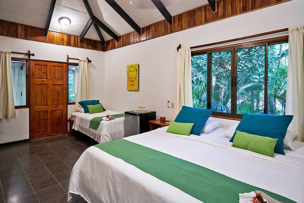Habitación de hotel con dos camas, una puerta de madera, una mesita con lavamanos y un ventanal con cortinas.