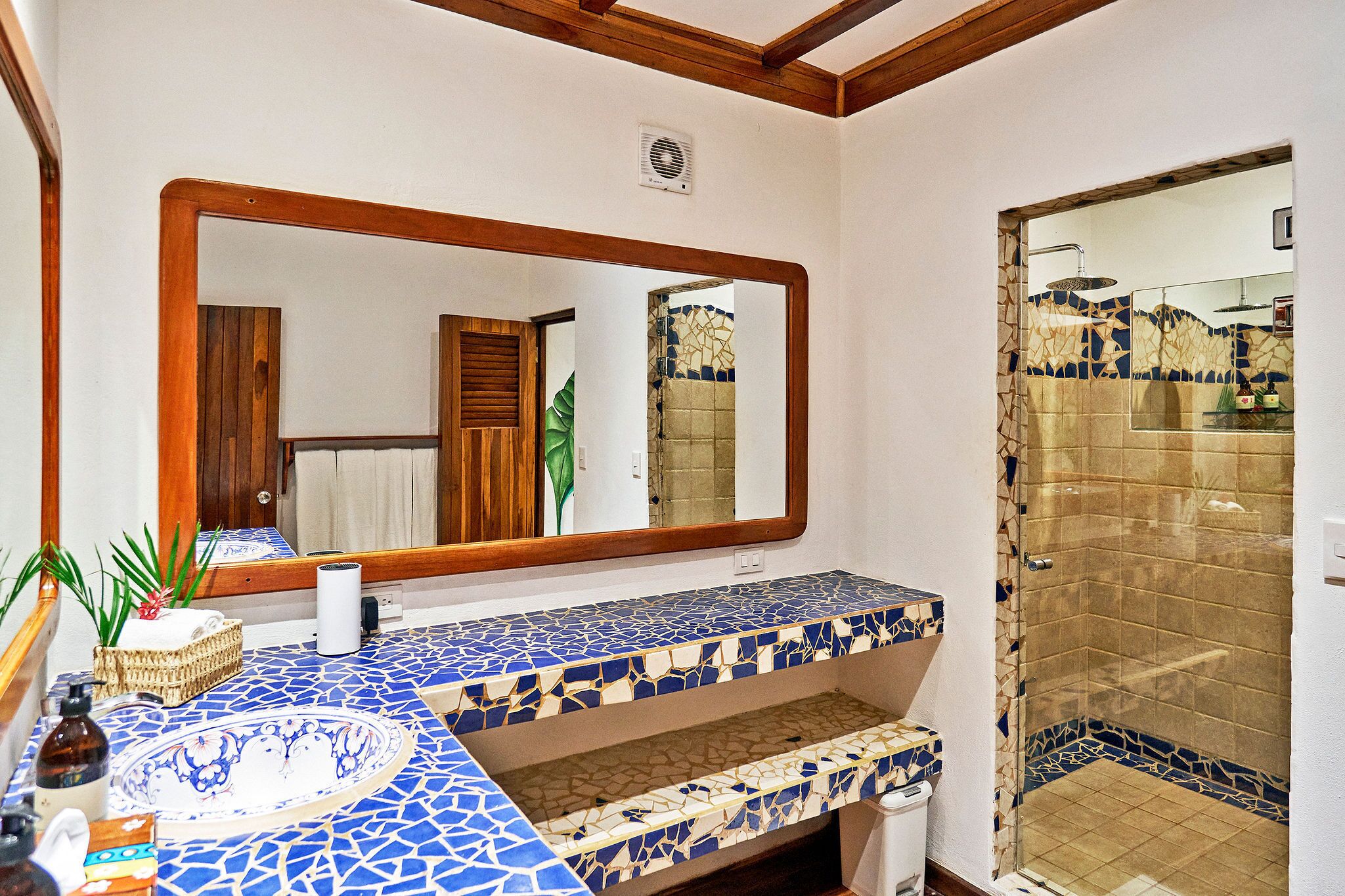 Une salle de bain dotée d’un grand miroir, d’un lavabo avec un plan de travail en mosaïque bleue et d’une douche séparée par une porte en verre.