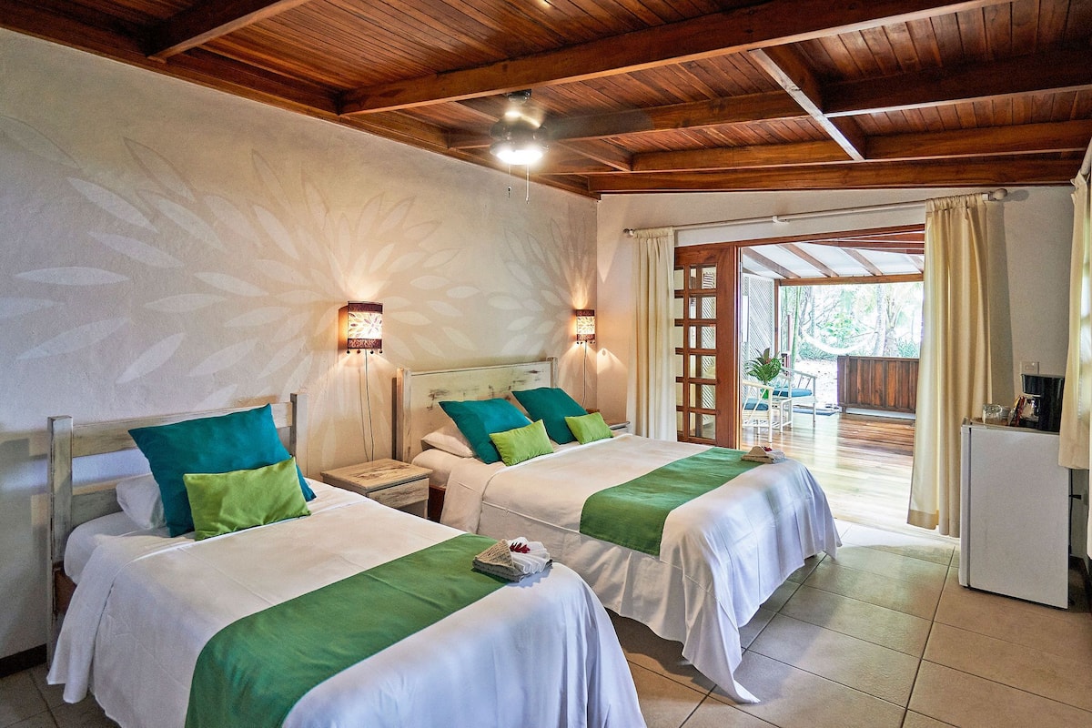 Habitación de hotel con dos camas, vigas de madera en el techo, lámparas fijadas a la pared y vistas al exterior.