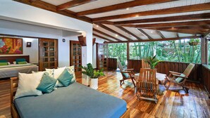 A spacious room with a large bed, wooden flooring, and a view of lush greenery through large windows. - Hotel Tropico Latino (Cóbano)