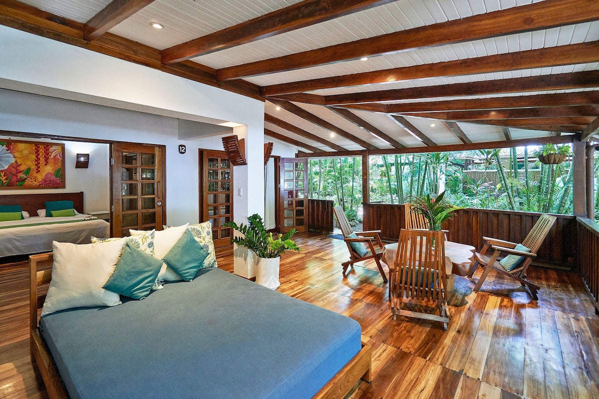 Una habitación amplia con una cama grande, suelo de madera y vistas a un follaje exuberante a través de amplios ventanales.