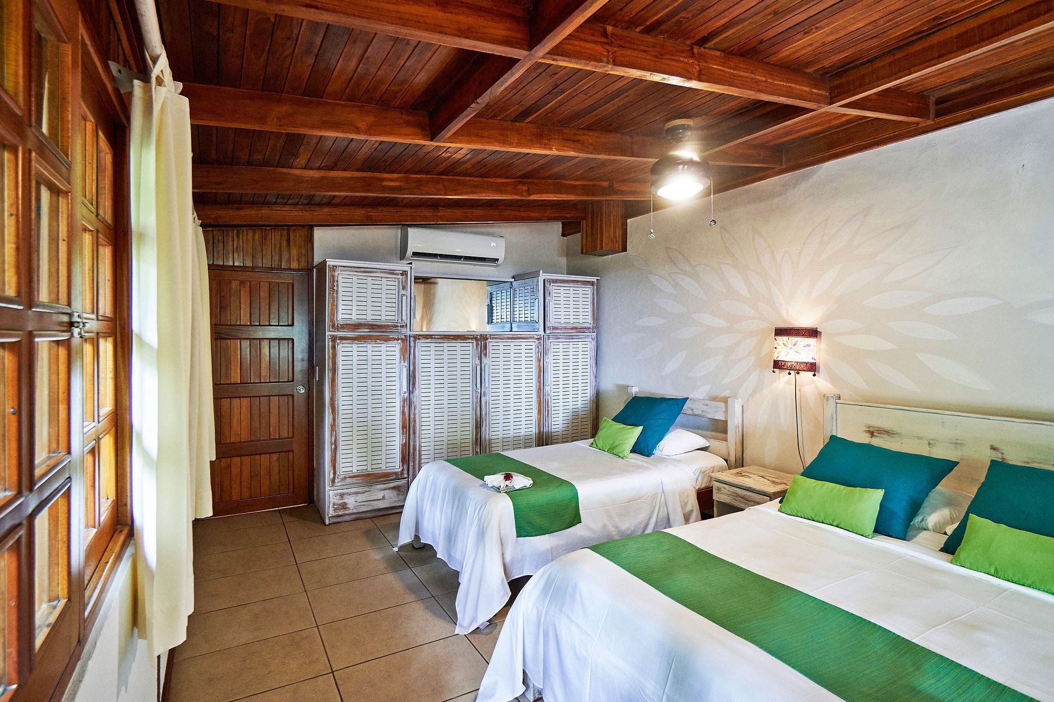 A hotel room with two beds, wooden ceiling beams, a ceiling fan, an air conditioning unit, and a wall-mounted light fixture.