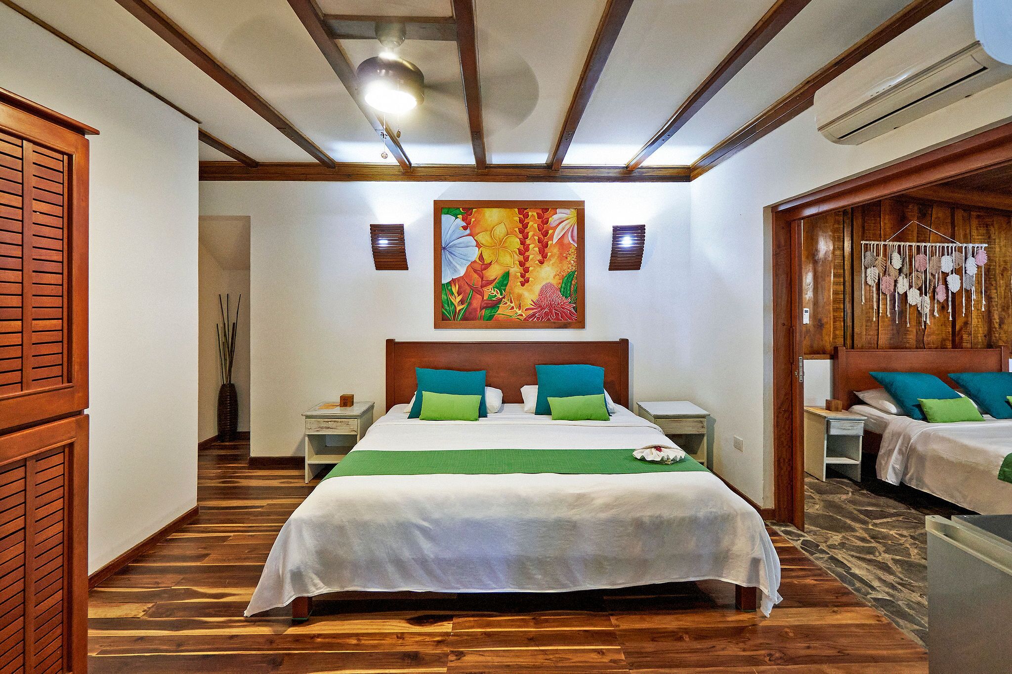 A hotel room with a large bed, two bedside tables, a nightstand, a wooden headboard, a ceiling fan, and a wall-mounted air conditioning unit.