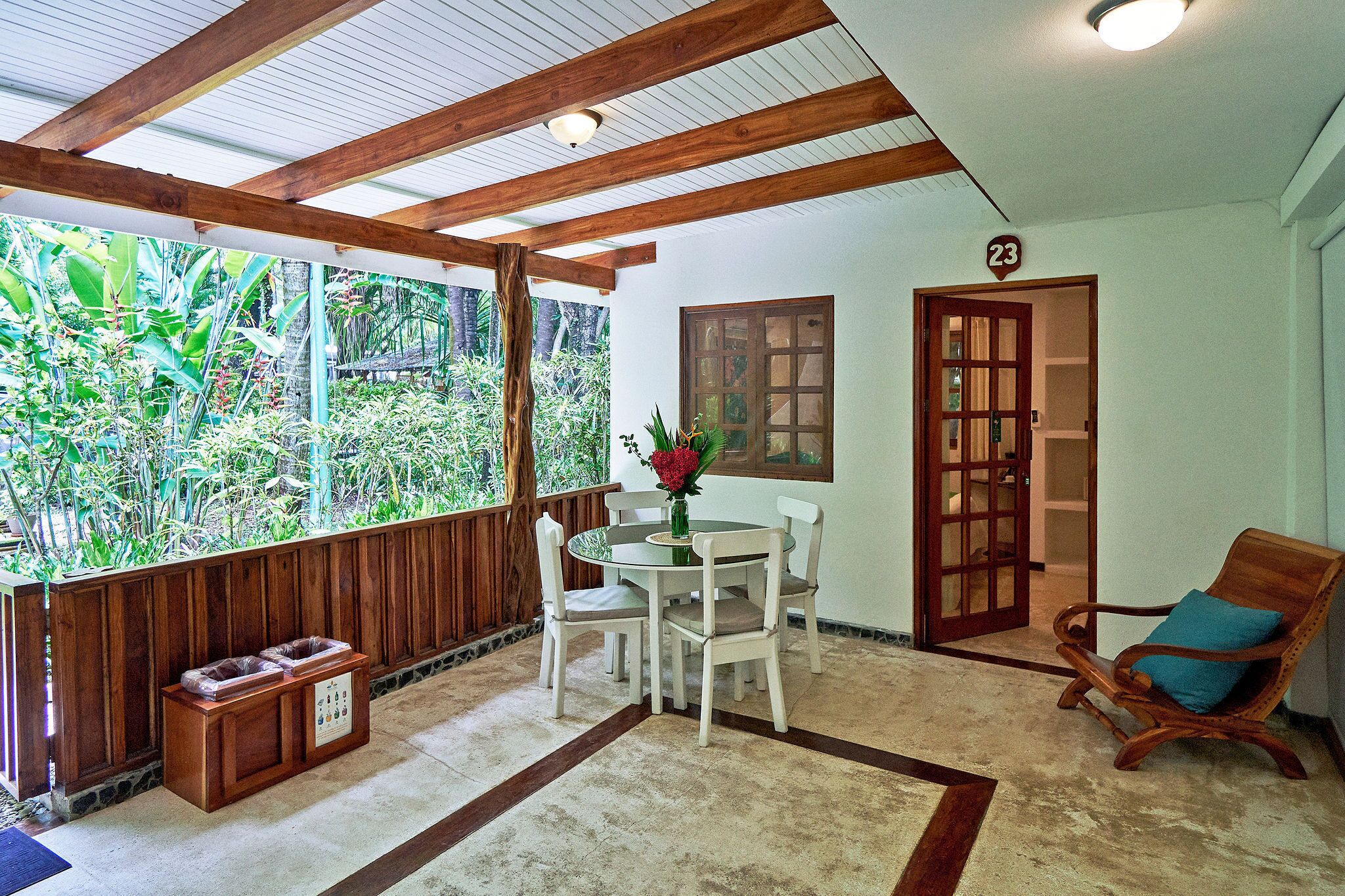 A room with a wooden ceiling, a round table with chairs, a wooden bench, a door with a number, and a view of greenery outside.