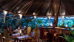 An outdoor restaurant with wooden tables and chairs, a bar area, and tropical plants in the background. - Hotel Tropico Latino (Cóbano)