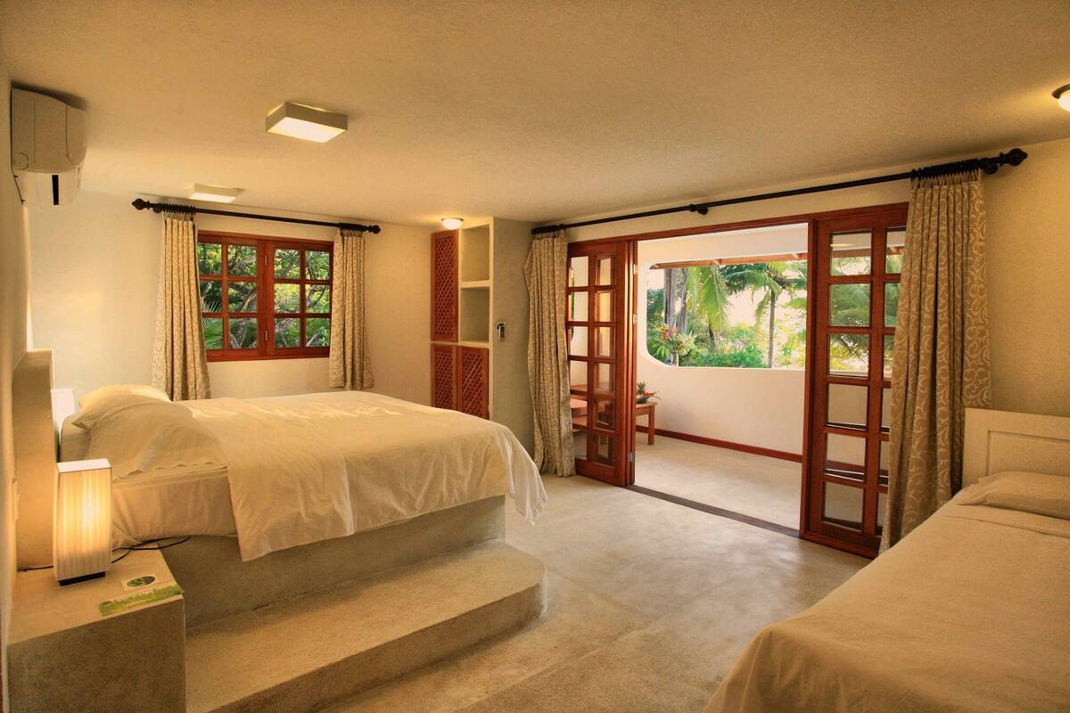 Habitación de hotel con dos camas, una mesita, una lámpara y vistas al exterior por una puerta.