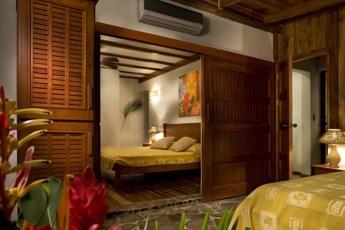 Una habitación con una cama, una puerta de madera, un aire acondicionado instalado en la pared y una mesita de noche con una lámpara.