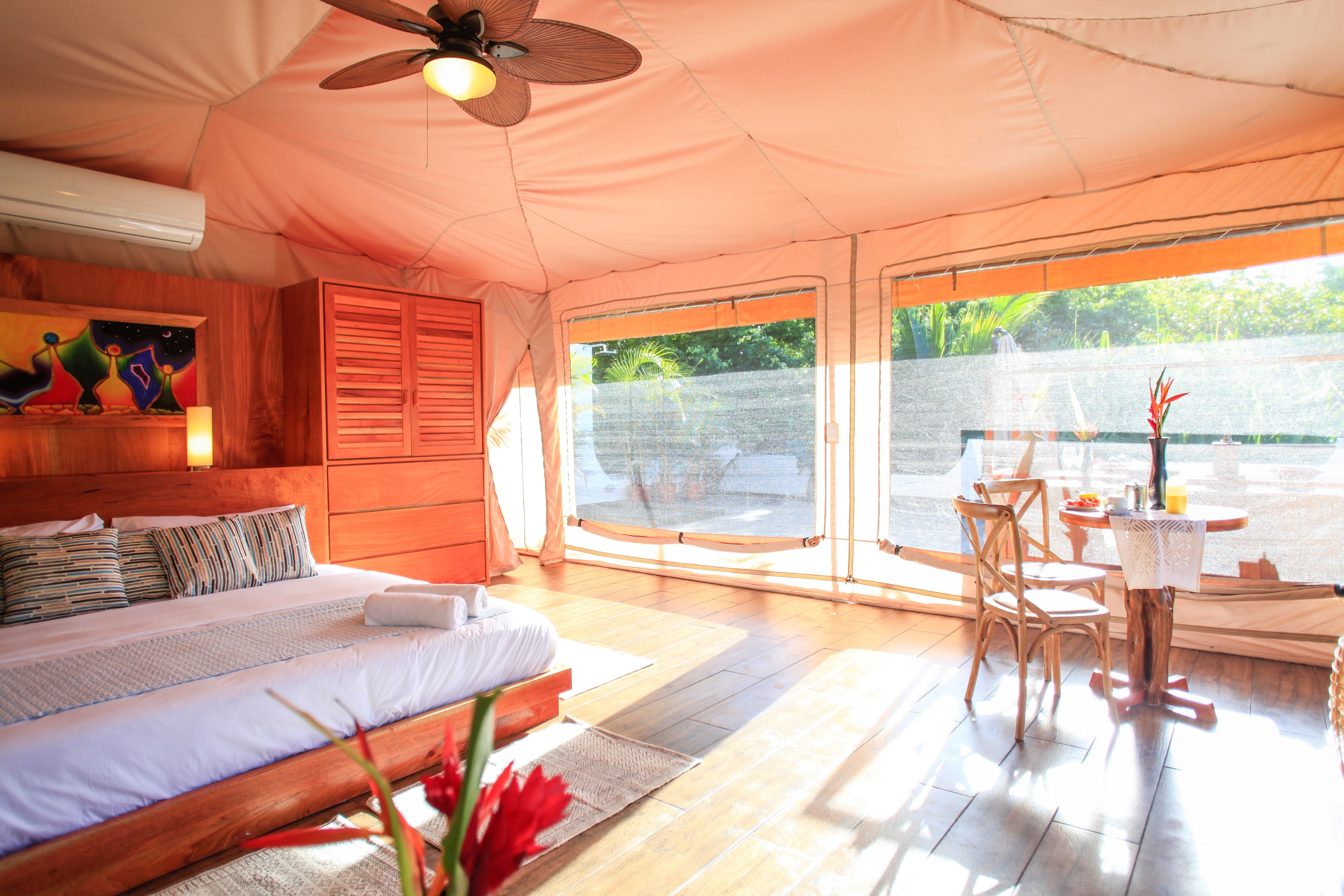 A spacious bedroom with a large bed, wooden headboard, and a ceiling fan. There is a window with a view of a garden and a balcony with a table and chairs.