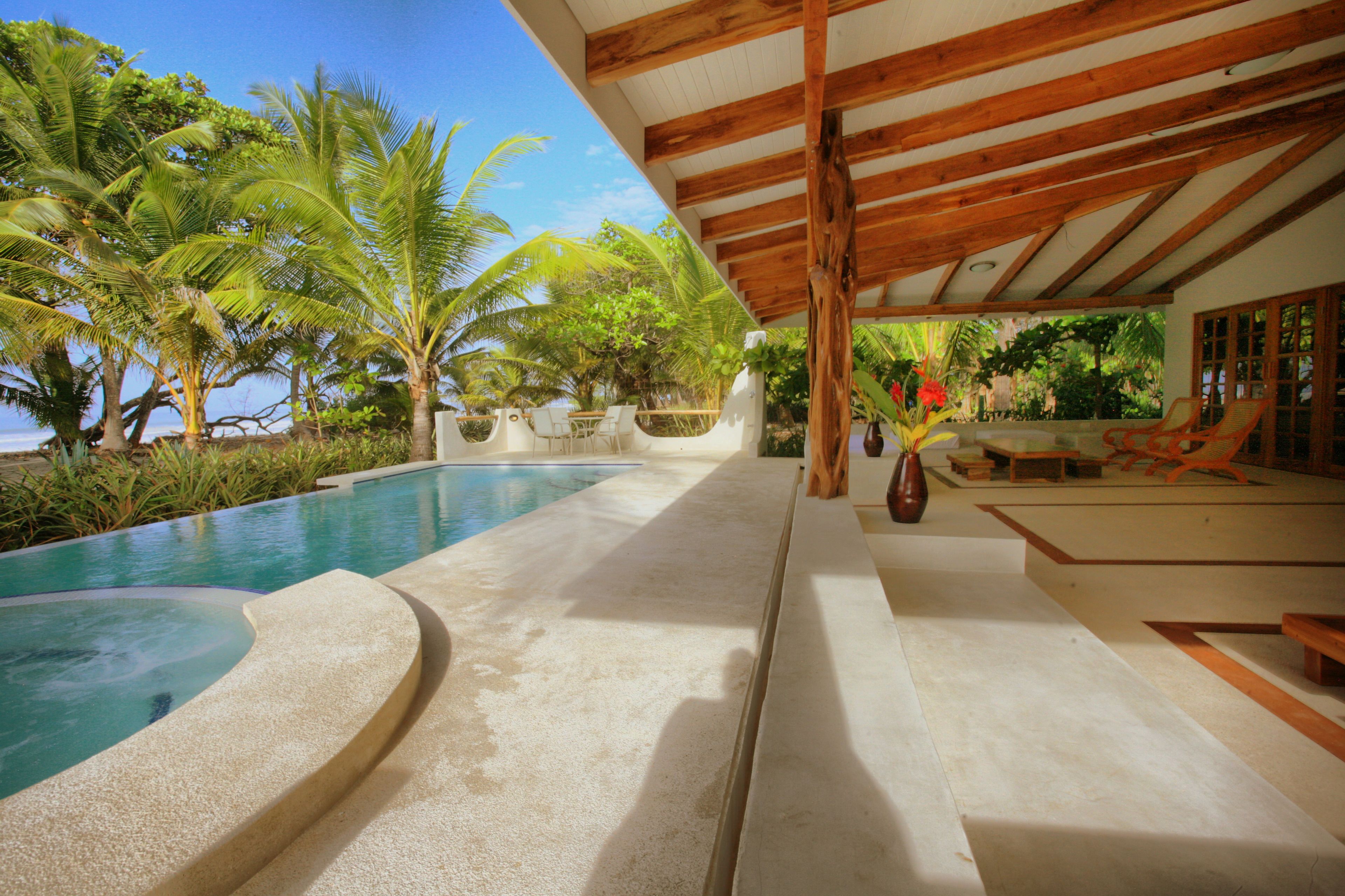 Un área de piscina con techo de madera, palmeras y un espacio de descanso con sillas.