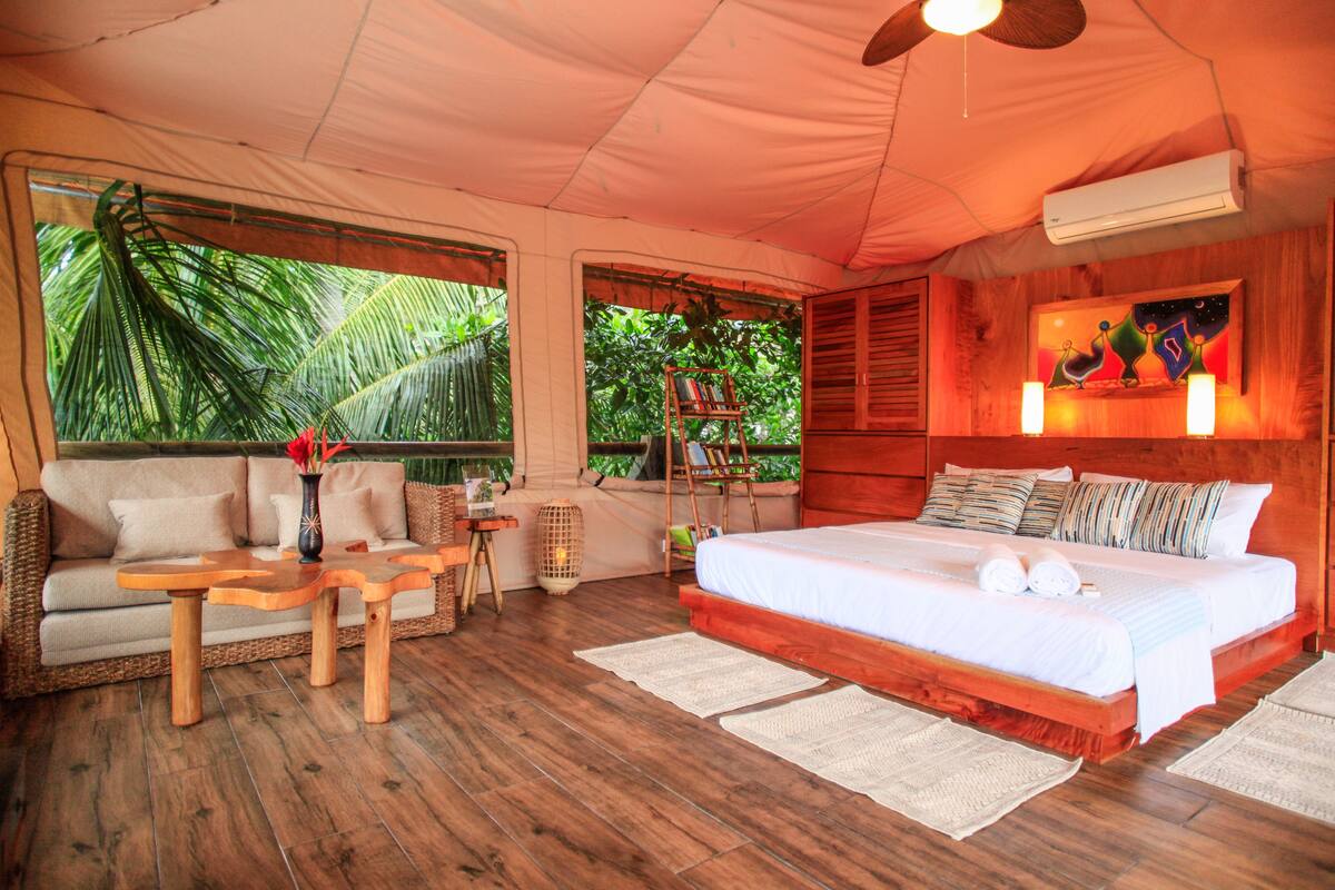 Habitación tipo tienda de campaña con cama, sofá, muebles de madera y ventanas grandes con vistas a la vegetación.