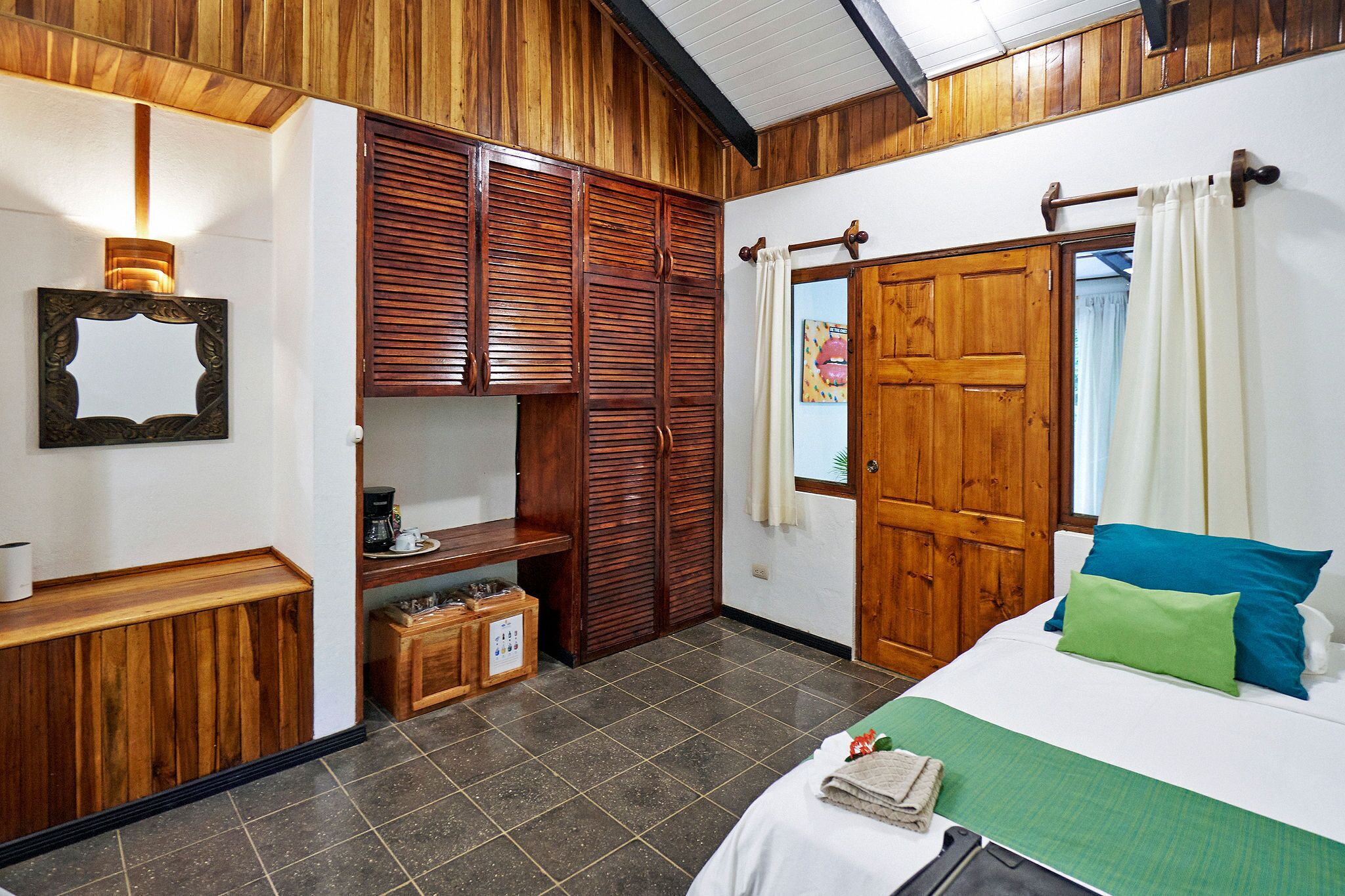 Quarto com cama, guarda-roupa de madeira, uma mesinha com cafeteira e chão ladrilhado.