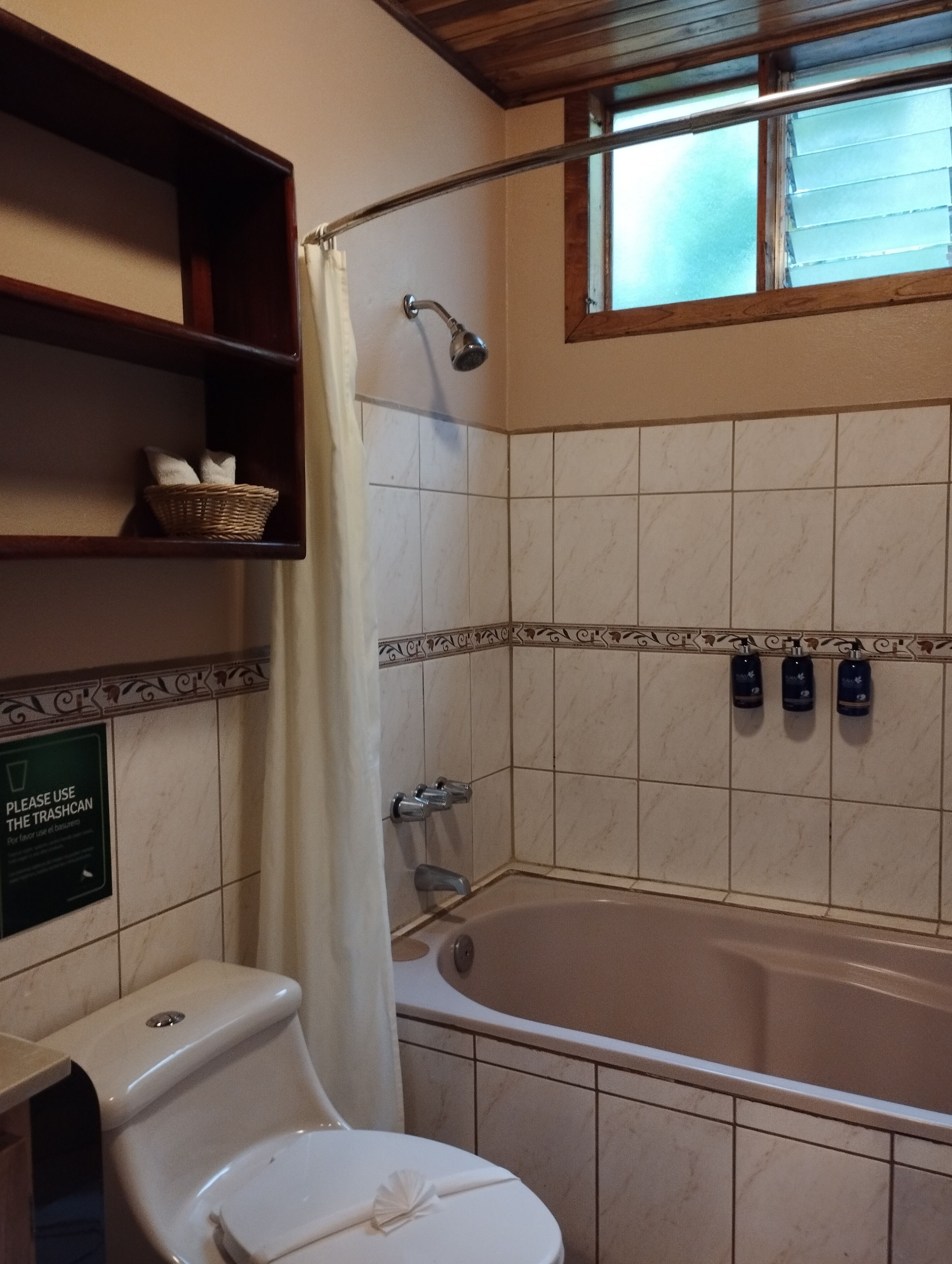 A bathroom with a bathtub, a toilet, and a shower curtain. There is a sign on the wall and a shelf with a basket.