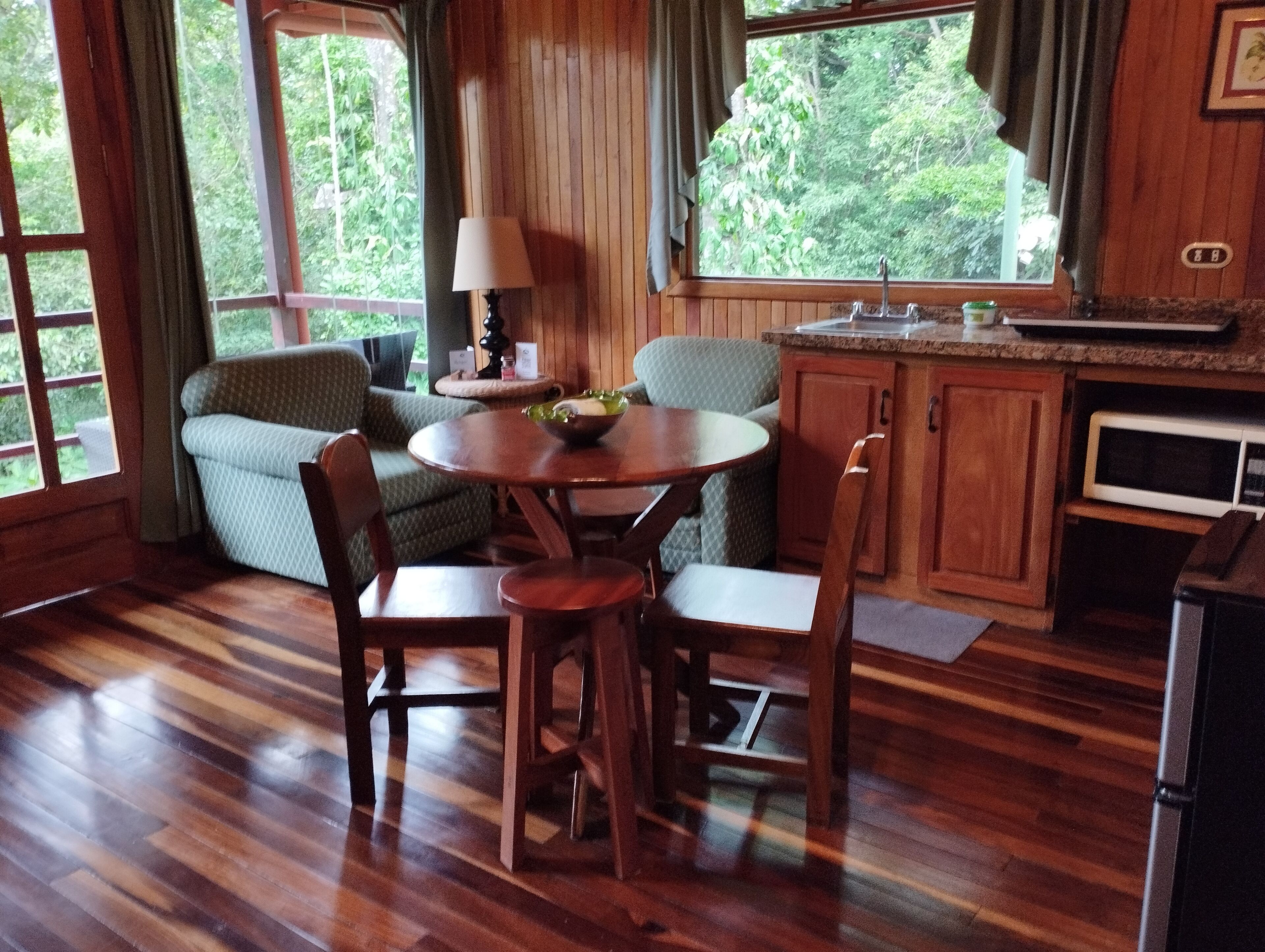 A cozy room with a wooden floor, a round wooden table with chairs, a green cushioned sofa, a small lamp, and a window with a view of greenery.