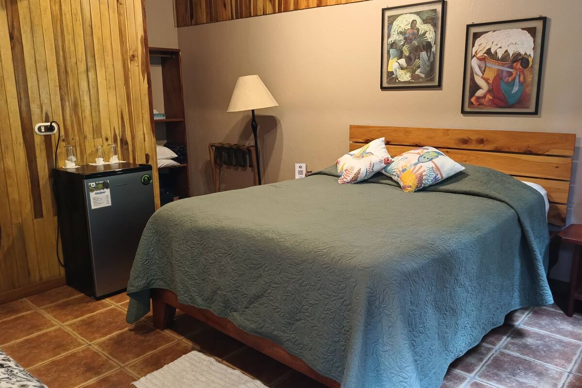 A bedroom with a bed, a refrigerator, a lamp, and two framed pictures on the wall.