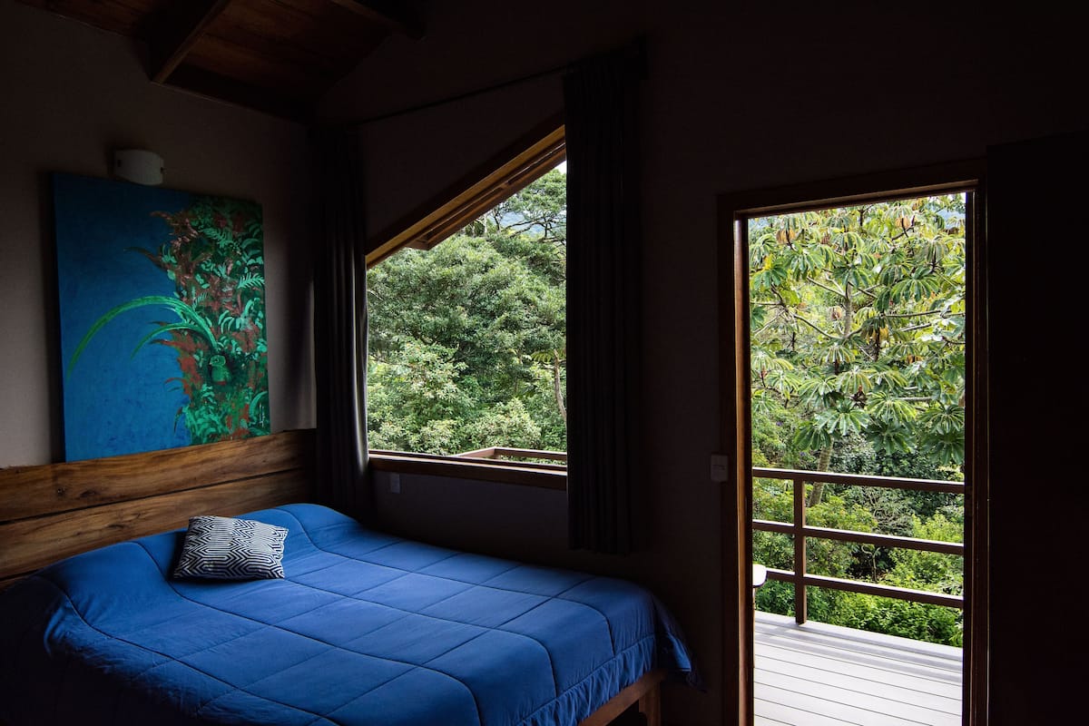 A bedroom with a bed, a painting, and a window with a view of trees.