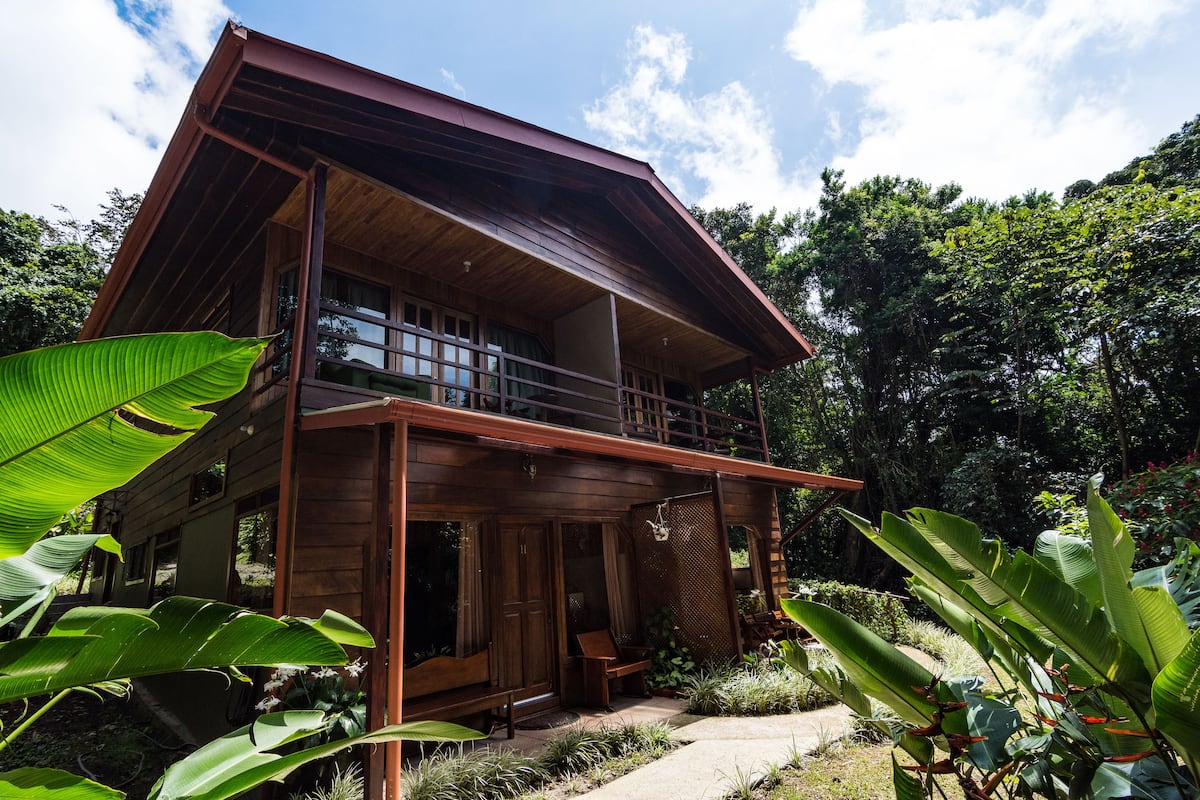 A two-story wooden house with a balcony, surrounded by lush greenery.