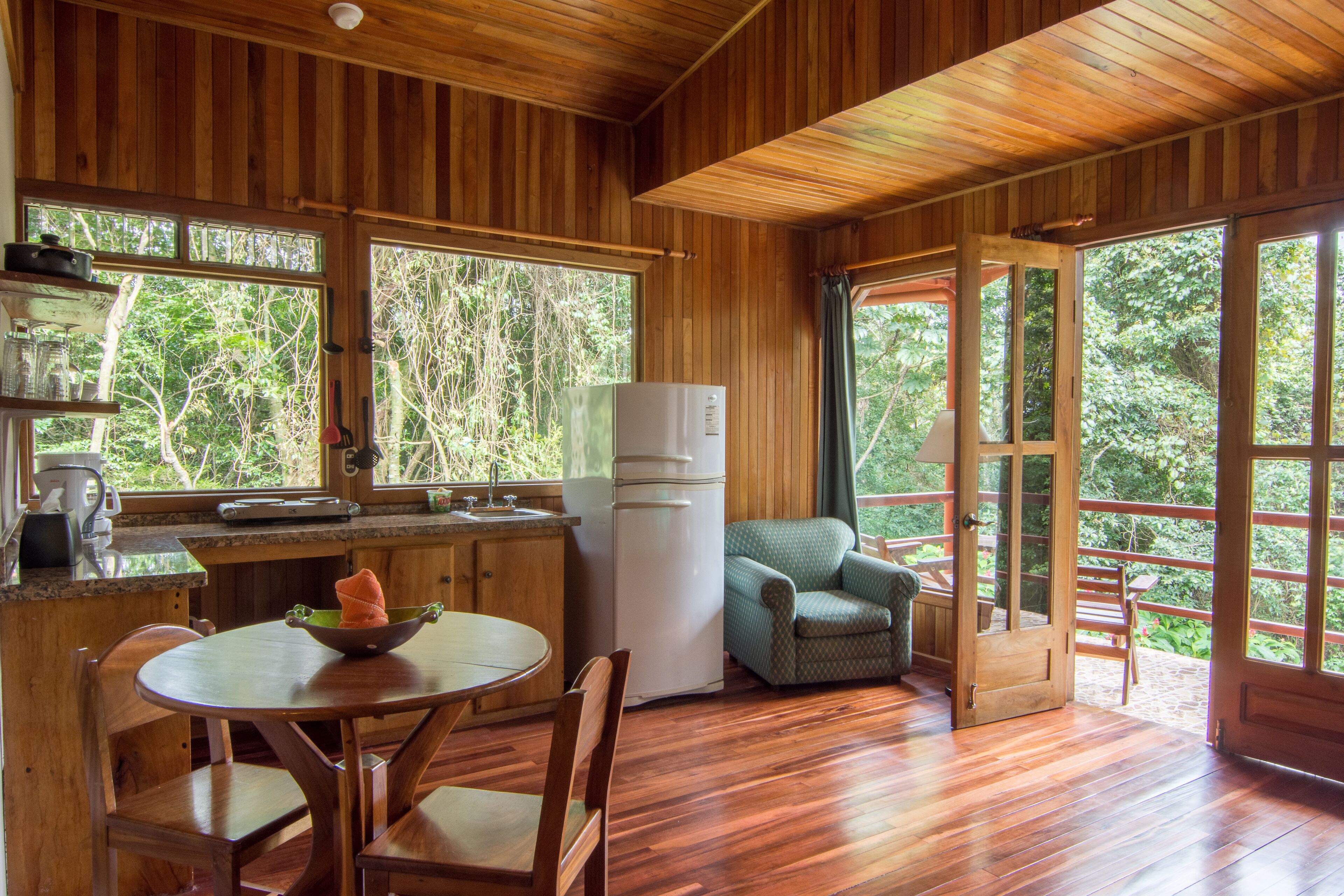 Une cabane en bois comprenant une cuisine, un coin repas et une grande fenêtre donnant sur une forêt.