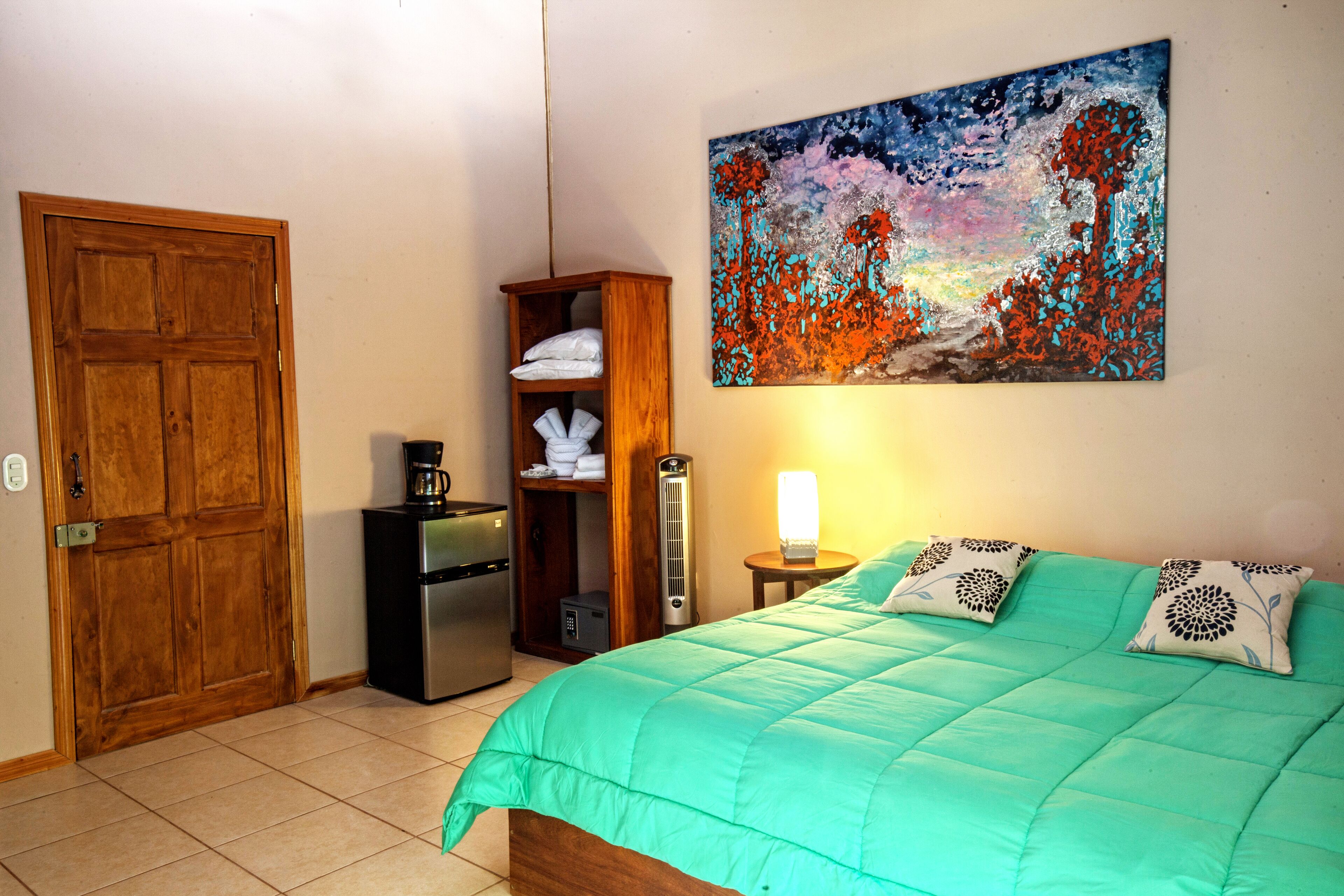 A bedroom with a green bedspread, a wooden wardrobe, a painting on the wall, and a bedside lamp.