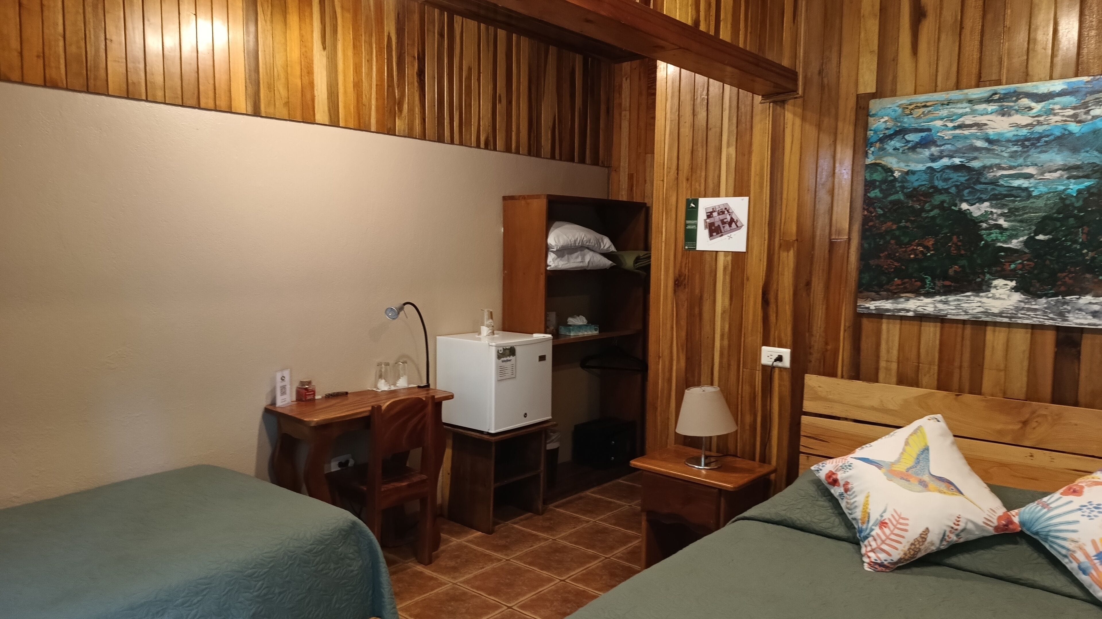 A cozy room with wooden paneling, a bed with a floral pillow, a desk with a lamp, a small refrigerator, and a painting on the wall.