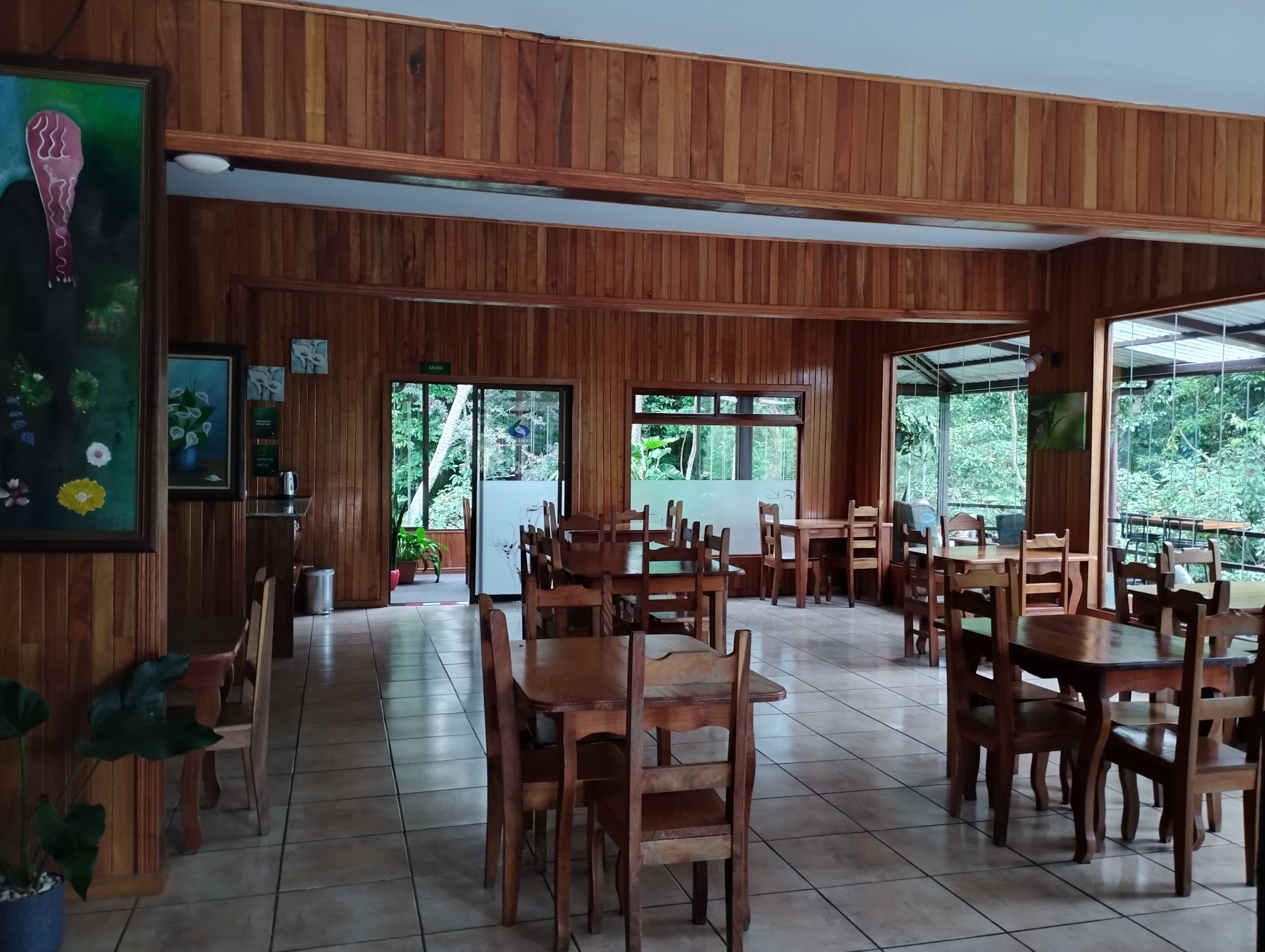 Una sala con paneles de madera, varias mesas y sillas, una puerta con la etiqueta 'SALON' y grandes ventanales que dan a zonas verdes.