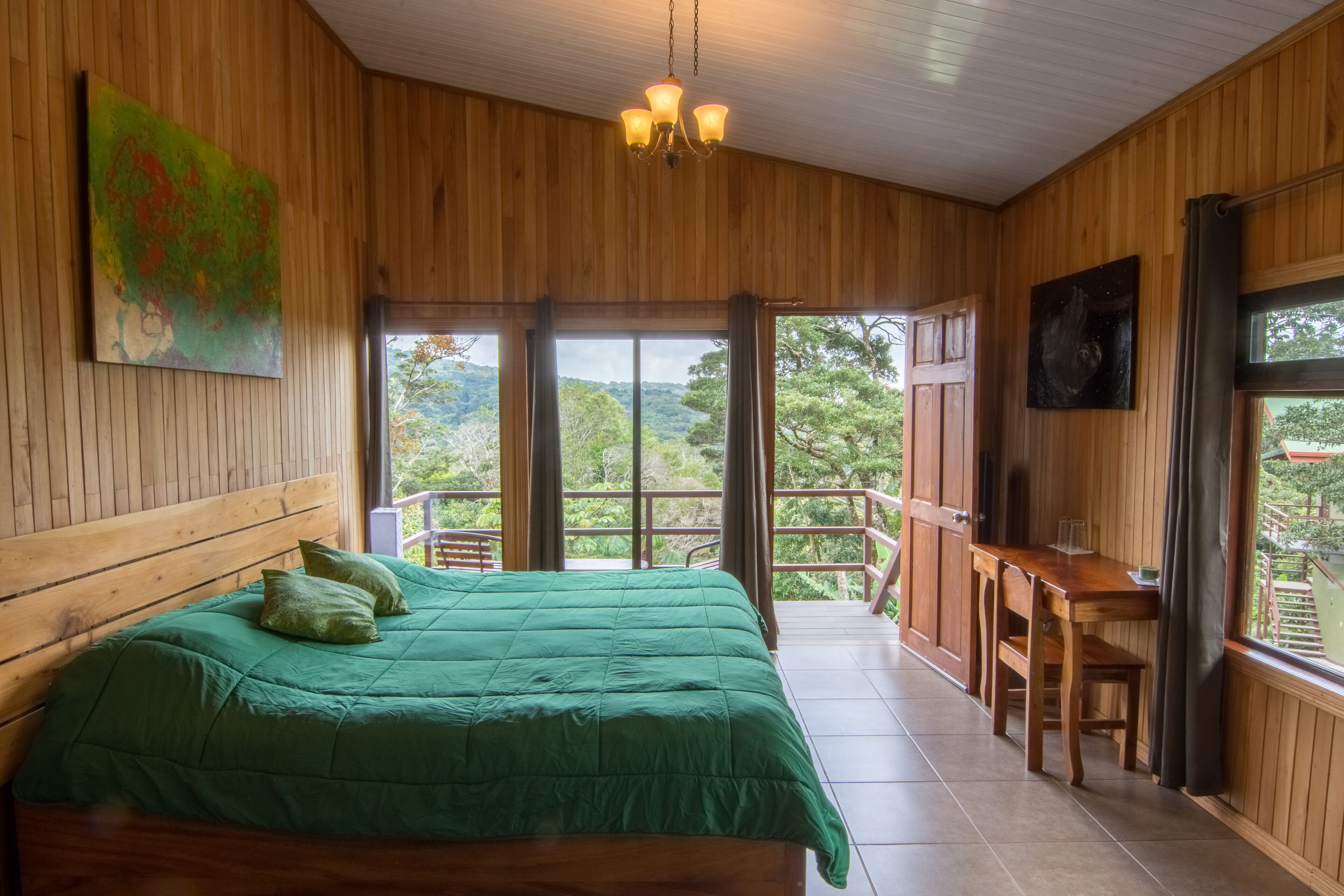 A wooden room with a bed, a desk, and a balcony with a view of the forest.