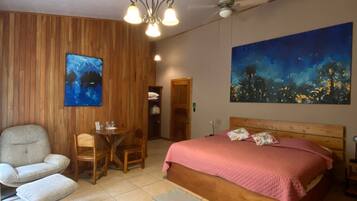 A bedroom with a bed, a chair, a table, a ceiling fan, and artwork on the walls.