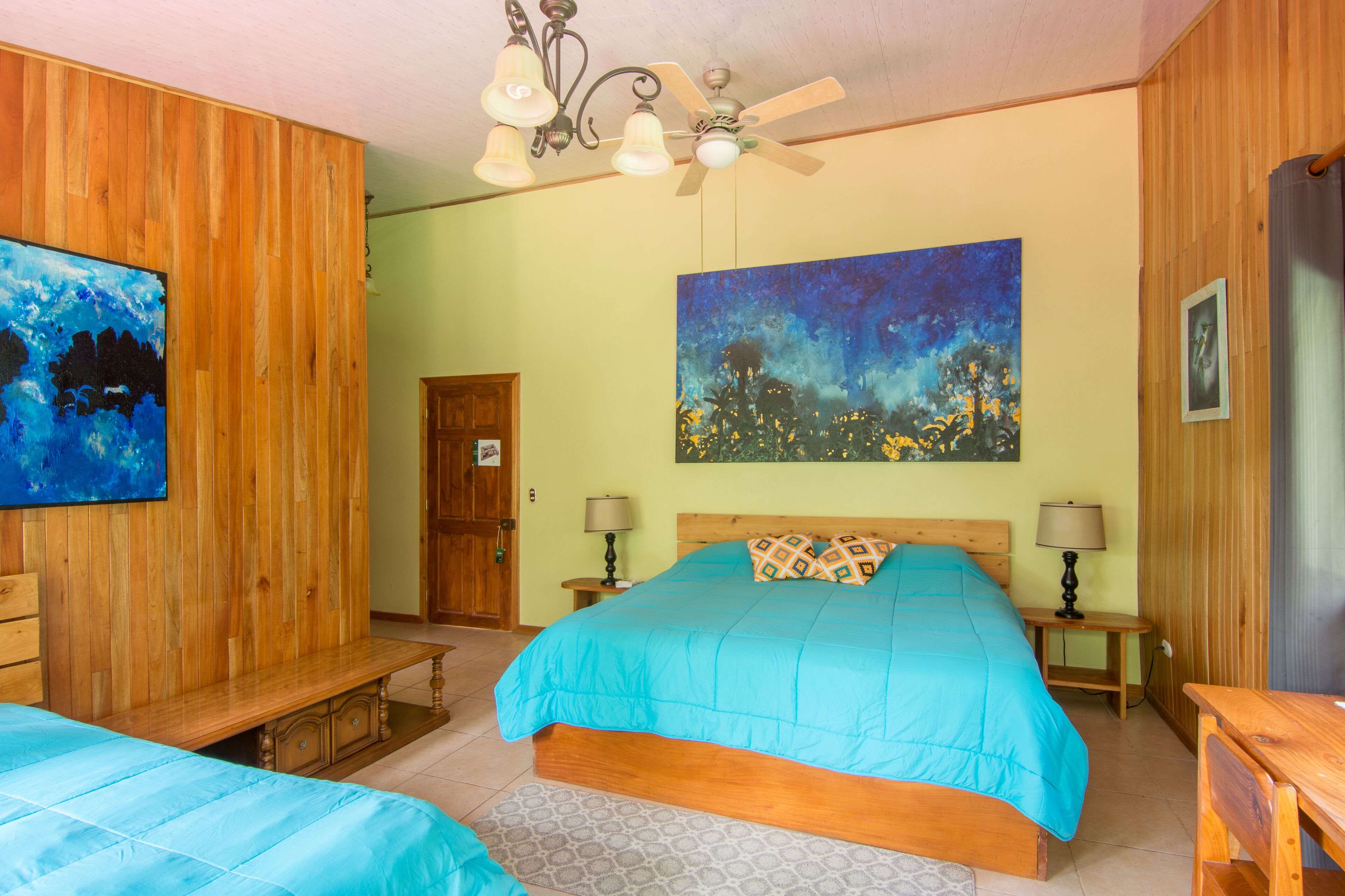 A bedroom with a wooden headboard, a large bed with a blue bedspread, two bedside tables with lamps, a wooden bench, and a painting on the wall.