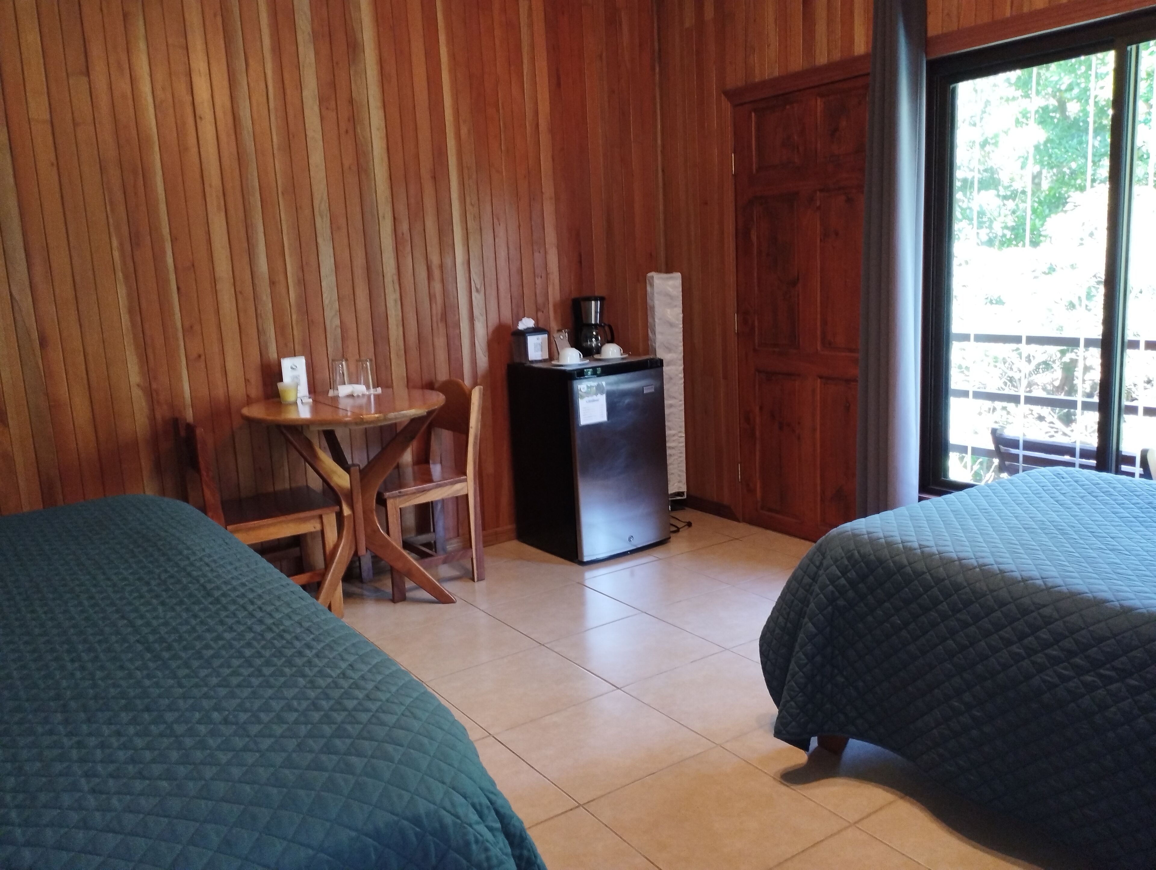 Una habitación con dos camas, una mesita con sillas, un refrigerador y una ventana con vistas a árboles.