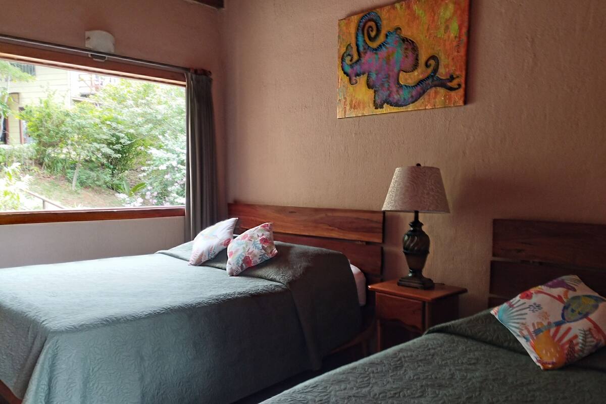 A hotel room with two beds, a window with a view of greenery, a painting on the wall, and a bedside lamp.