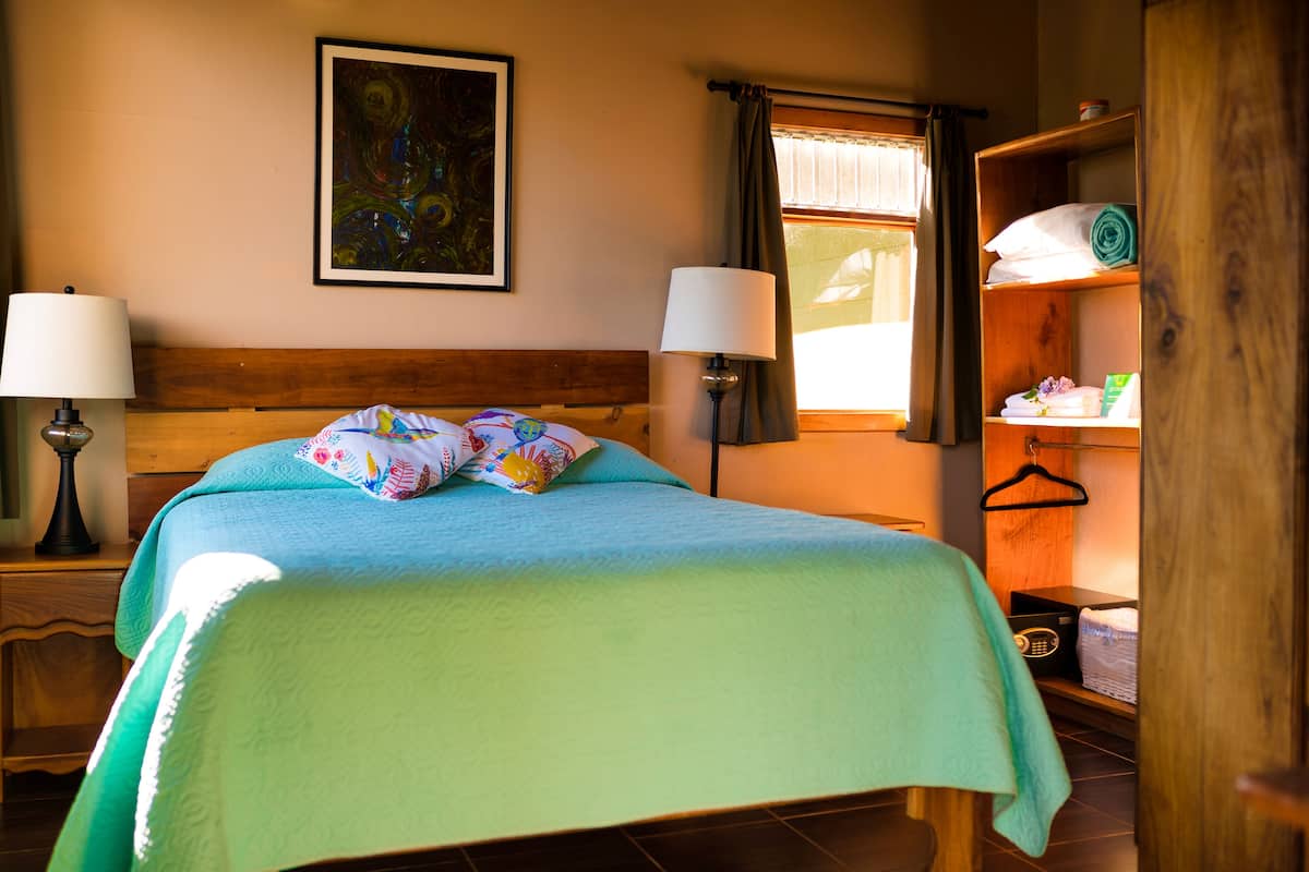 A neatly made bed with a blue bedspread and pillows, two bedside lamps, a wooden headboard, a painting on the wall, and a window with curtains.